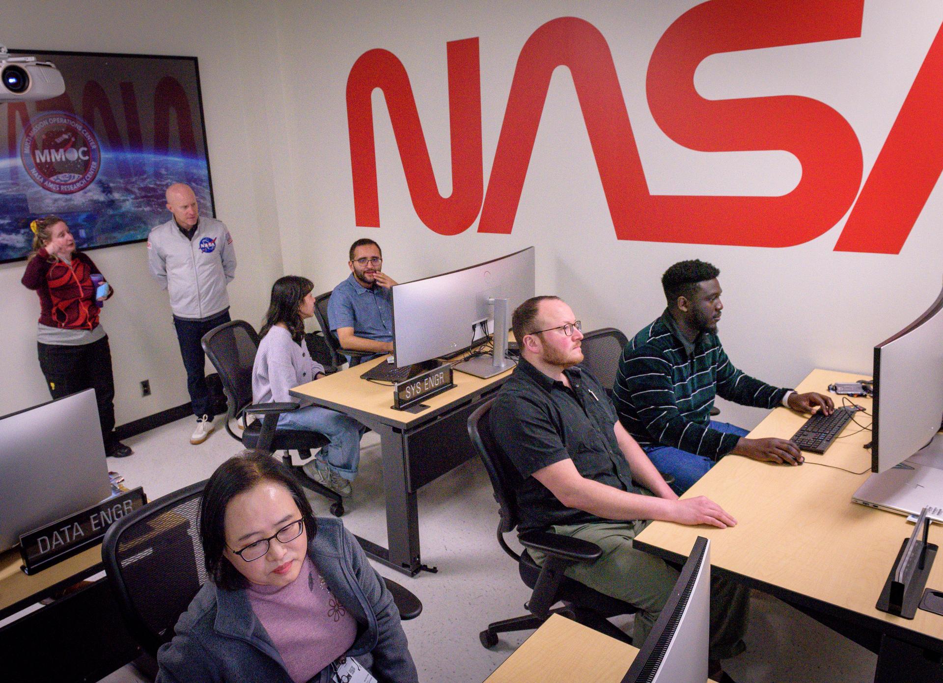 TechEdSat-11 operators Heather Smith, left, Justin Pane, Daphne Dao and Alejandro Salas, seated, Kyeong Ja Kim, Luke Idziak, and Kwabena Boateng right, in the Ames Multi-Mission Operations Center (MMOC), N240 Annex, Eros control room 162. The team monitors the spacecraft's status during the Exo-Brake “parachute” deployment.   The ExoBrake is a drag device that increases the total surface area of the spacecraft to assist with a quicker deorbit. This maneuver is deployed at the end of mission to satisfy NASA's deorbit requirement and prevent space debris. TechEdSat has spent the last several months coordinating with NASA’s Conjunction Assessment Risk Analysis (CARA) team to ensure the spacecraft can safely deploy the ExoBrake without colliding into any other objects.