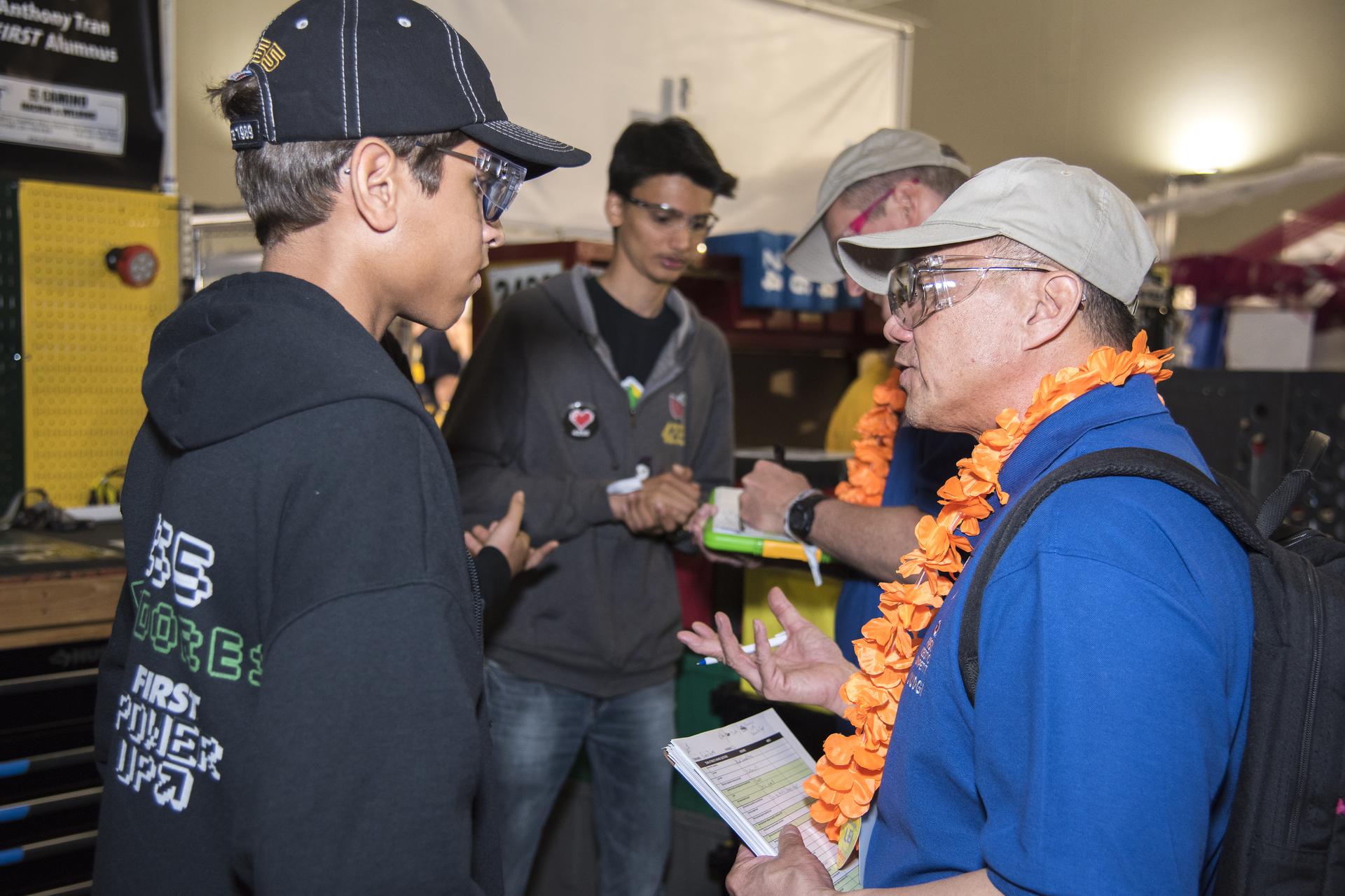 NASA systems engineer, right, in a bright blue shirt, tan baseball cap, and orange flowered lei, talks with a student participant from the Aerospace Valley Robotics Competition who is wearing a black hooded sweatshirt and a black baseball cap. In the background, another student in a gray, zip-up sweatshirt speaks with another NASA judge.