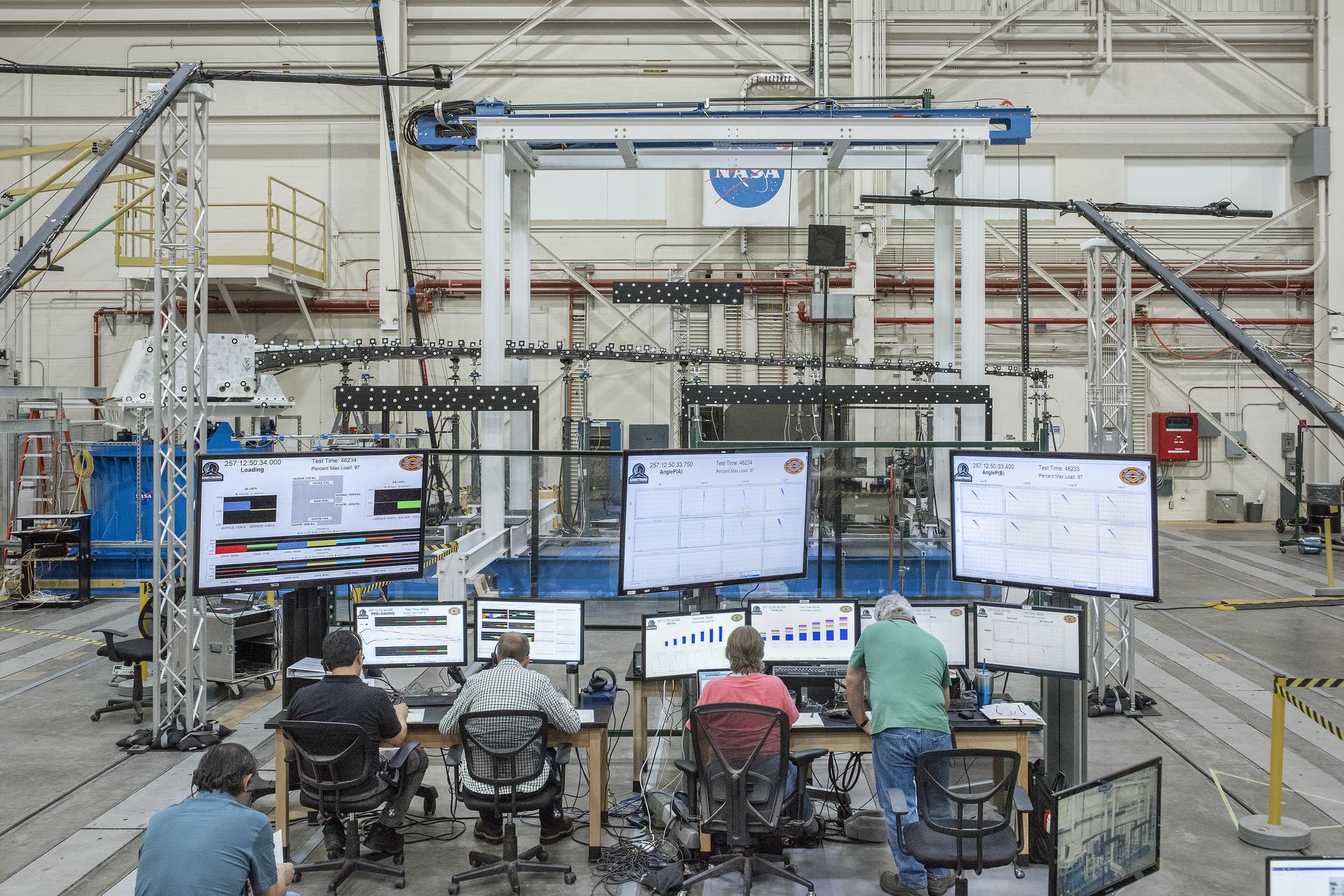 Broad view of five people sitting and standing behind nine computer screens during loads testing of an aircraft wing. The test setup is full of structures and wires surrounding the test article.