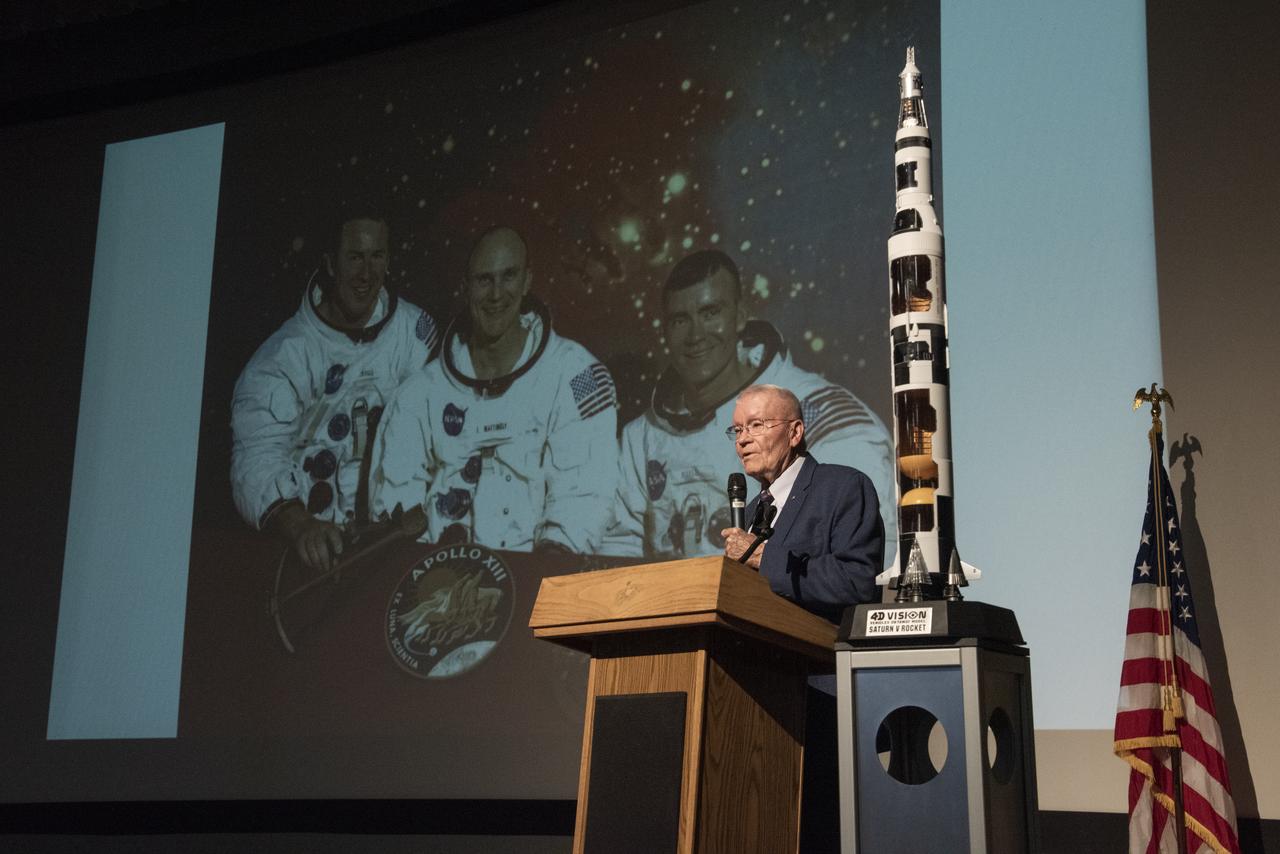 Apollo Astronaut Fred Haise speaks to a crowd of NASA and U.S Air Force employees at the Edwards Air Force Base theater about his career with NASA and as a military pilot. Haise stands on stage with a photo of former astronauts Jim Lovell and Jack Swigert who accompanied him on the Apollo 13 lunar mission in the background with a model of the Saturn V rocket.