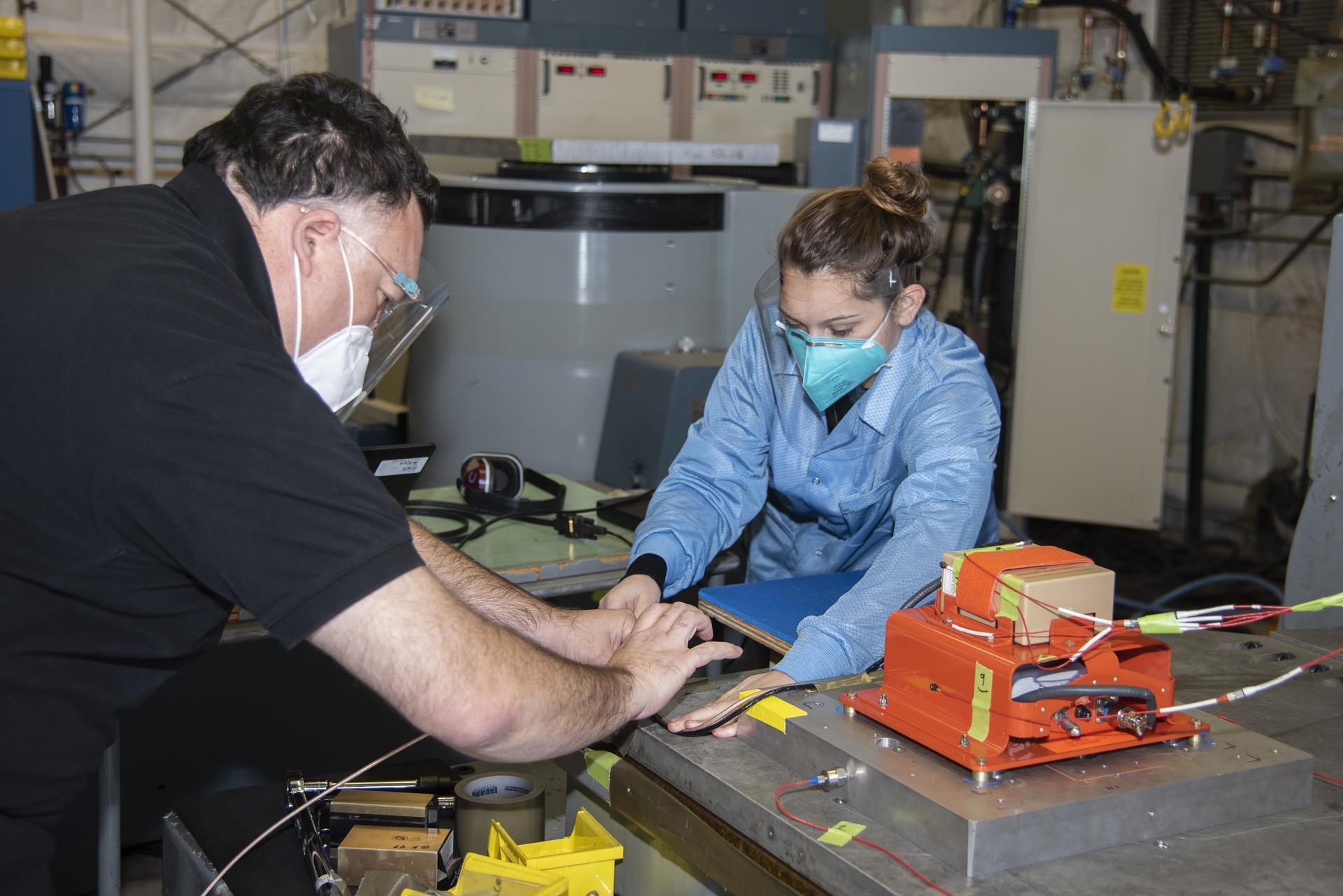 Woman working with a man on a piece of equipment used for testing. They both wear safety goggles and masks as they manipulate the instrument component with their hands. The woman wears a blue jacket while the man wears a short-sleeve black polo.