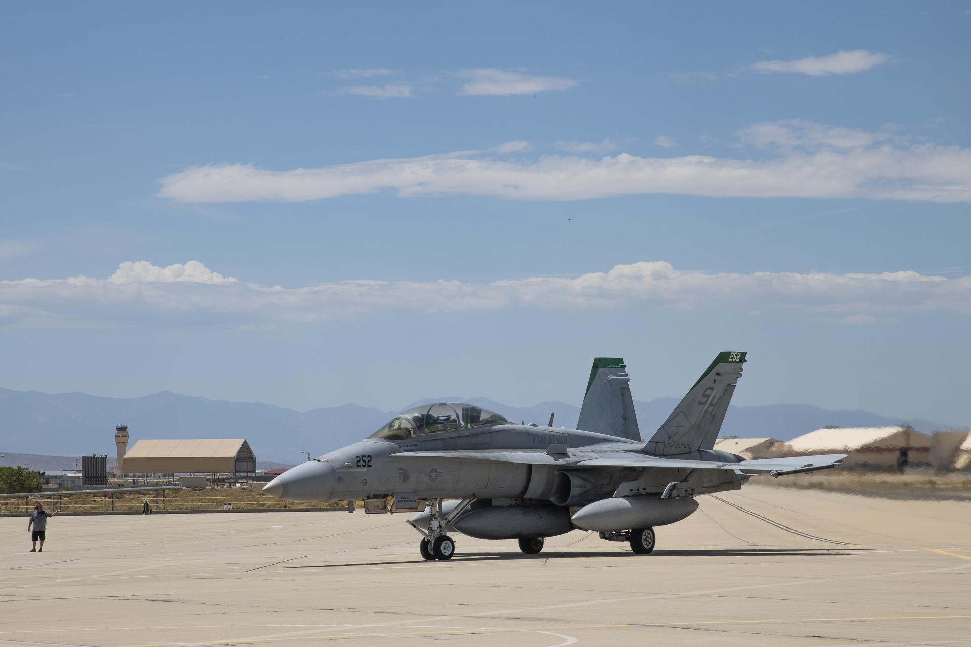 A person is standing, motioning toward an aircraft as it arrives at Armstrong. There are tan buildings in the distant background.