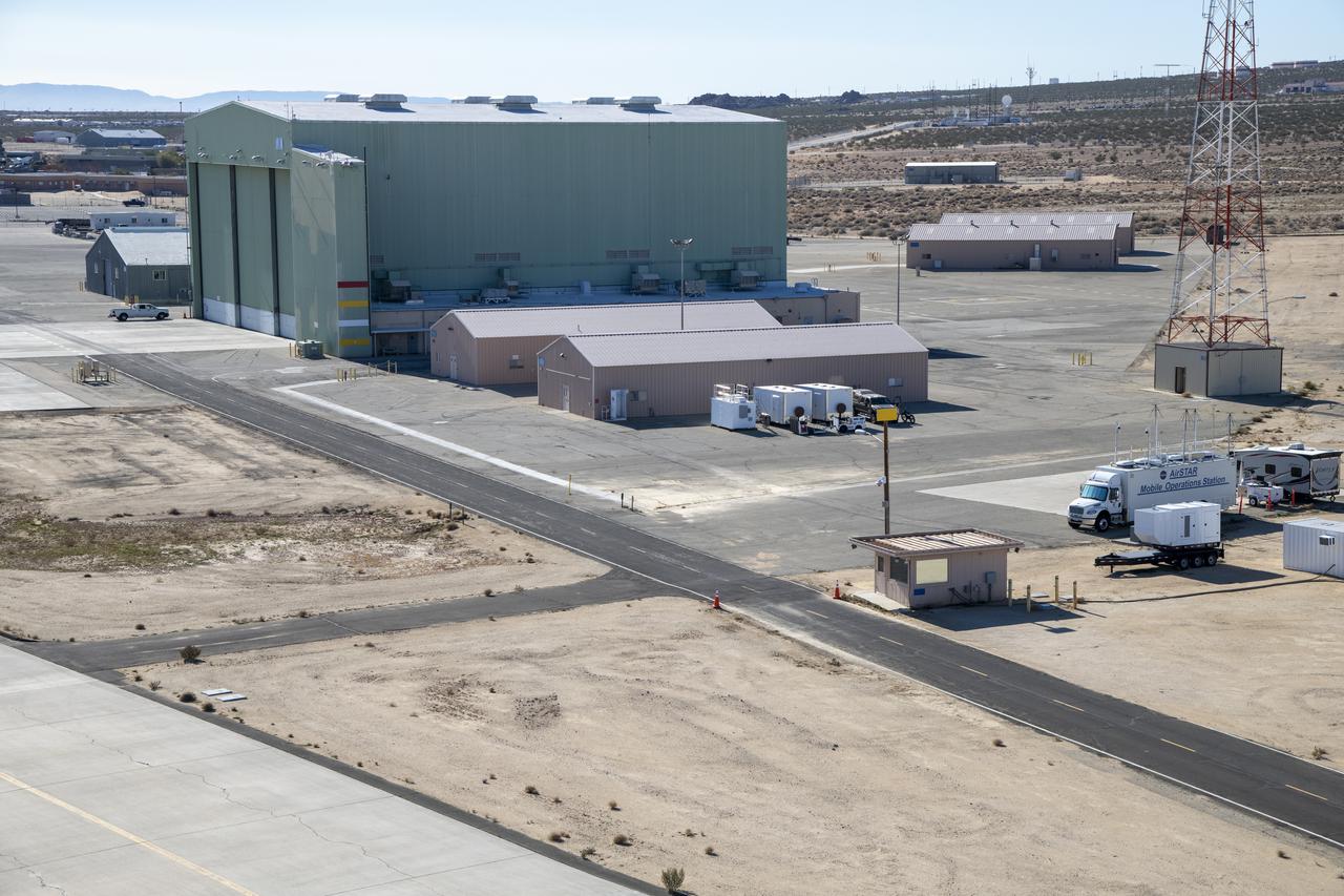 An aerial image taken by one of NASA’s photographers during recent helicopter flights shows a view of the building 4833 structure and the mobile operating facility at NASA’s Armstrong Flight Research Center in Edwards, California. NASA’s Advanced Air Mobility National Campaign uses the mobile operations facility vehicle shown in the lower right corner during test operations. The red, yellow, and white building markings applied to building 4833 are used to provide visual aids to the pilot during handling qualities testing used to research advanced air mobility flight requirements.