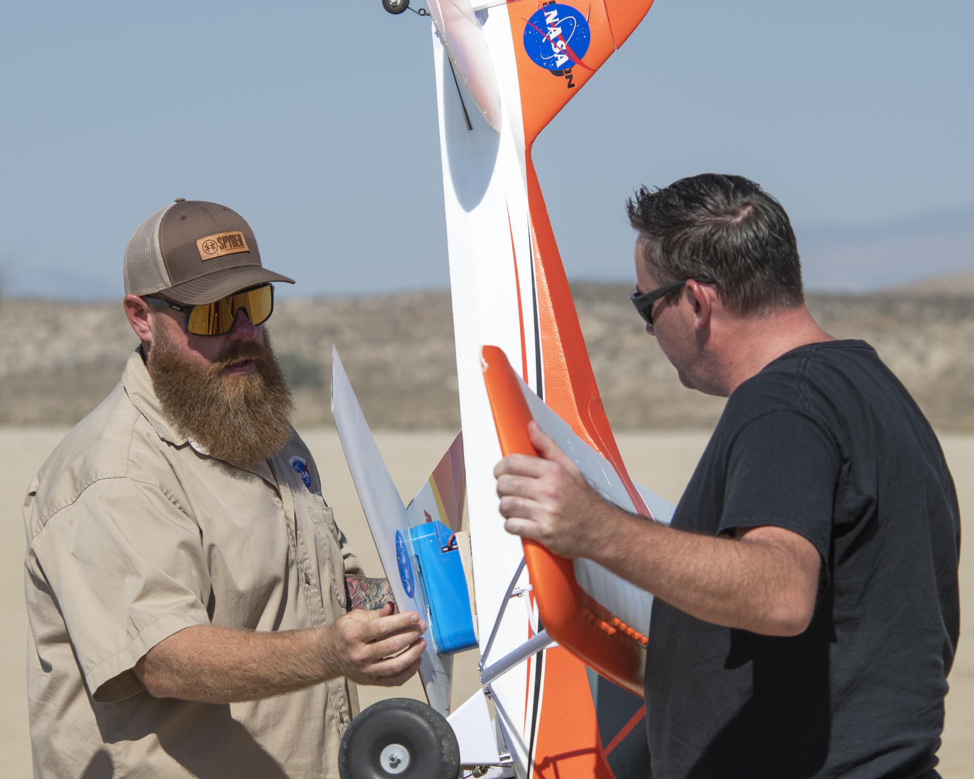 One man holds a large remotely piloted aircraft, while another man attaches a smaller aircraft to it.