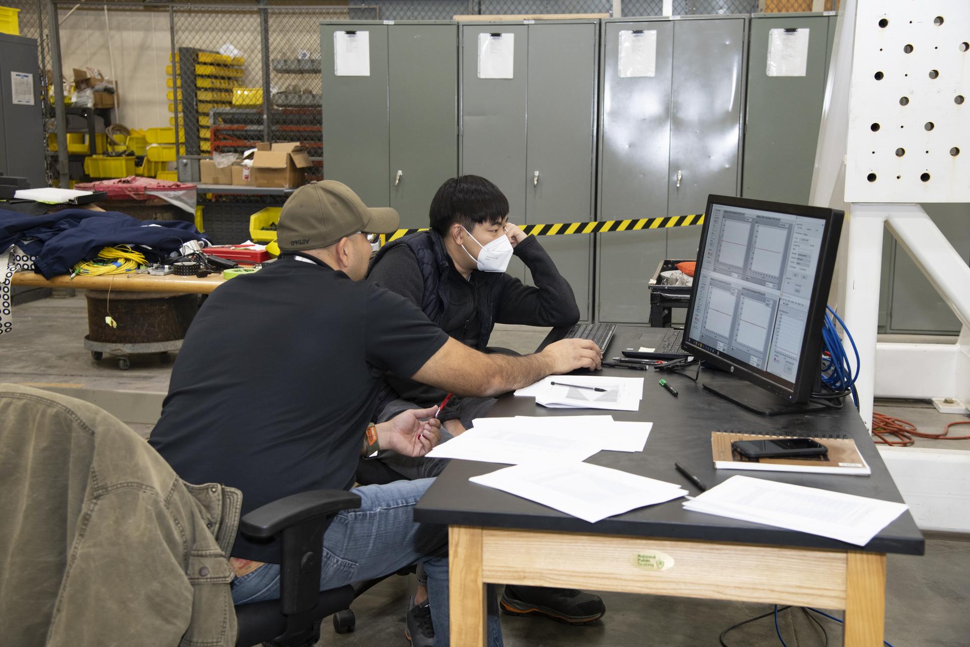 Two people site at a desk in front of a computer screen. The person sitting behind and to the right of the other person is wearing a white mask. In the background is a row of four to five gray lockers.