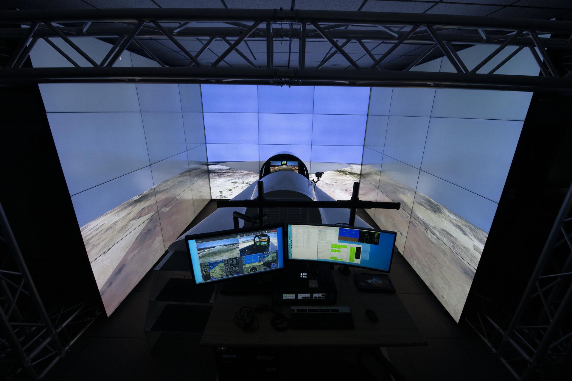 Interior view of the X-59 flight simulator at NASA’s Armstrong Flight Research Center in Edwards, California, showing a fully equipped cockpit with wraparound high-resolution display screens and control panels. The simulator replicates the layout of the X-59 aircraft and is used by test pilots to train for upcoming Quesst missions aimed at reducing sonic booms to quiet sonic "thumps."