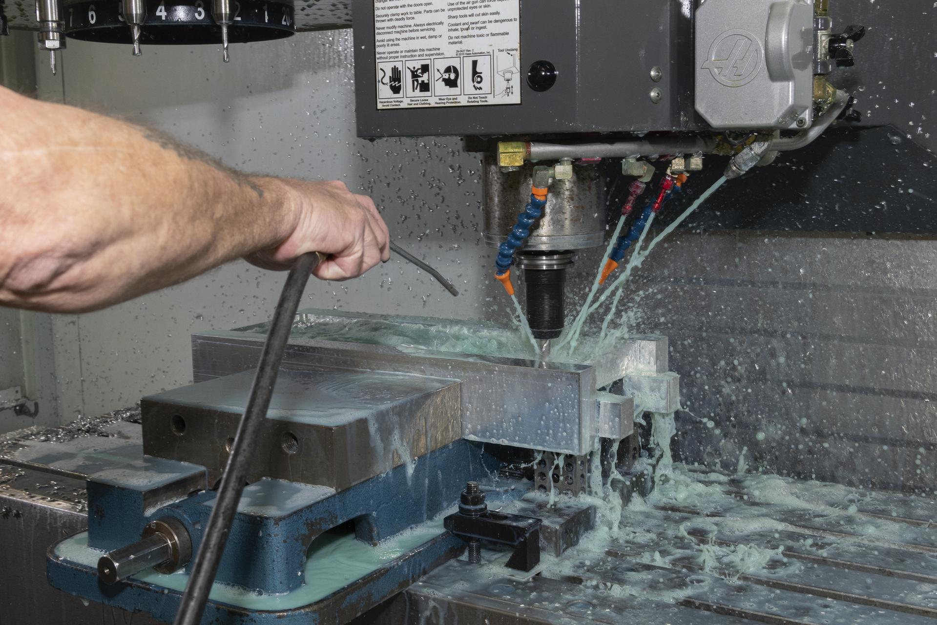 A technician is using a machine to drill pieces of silver metal; there is a light blue liquid coming out of jets next to the drill.