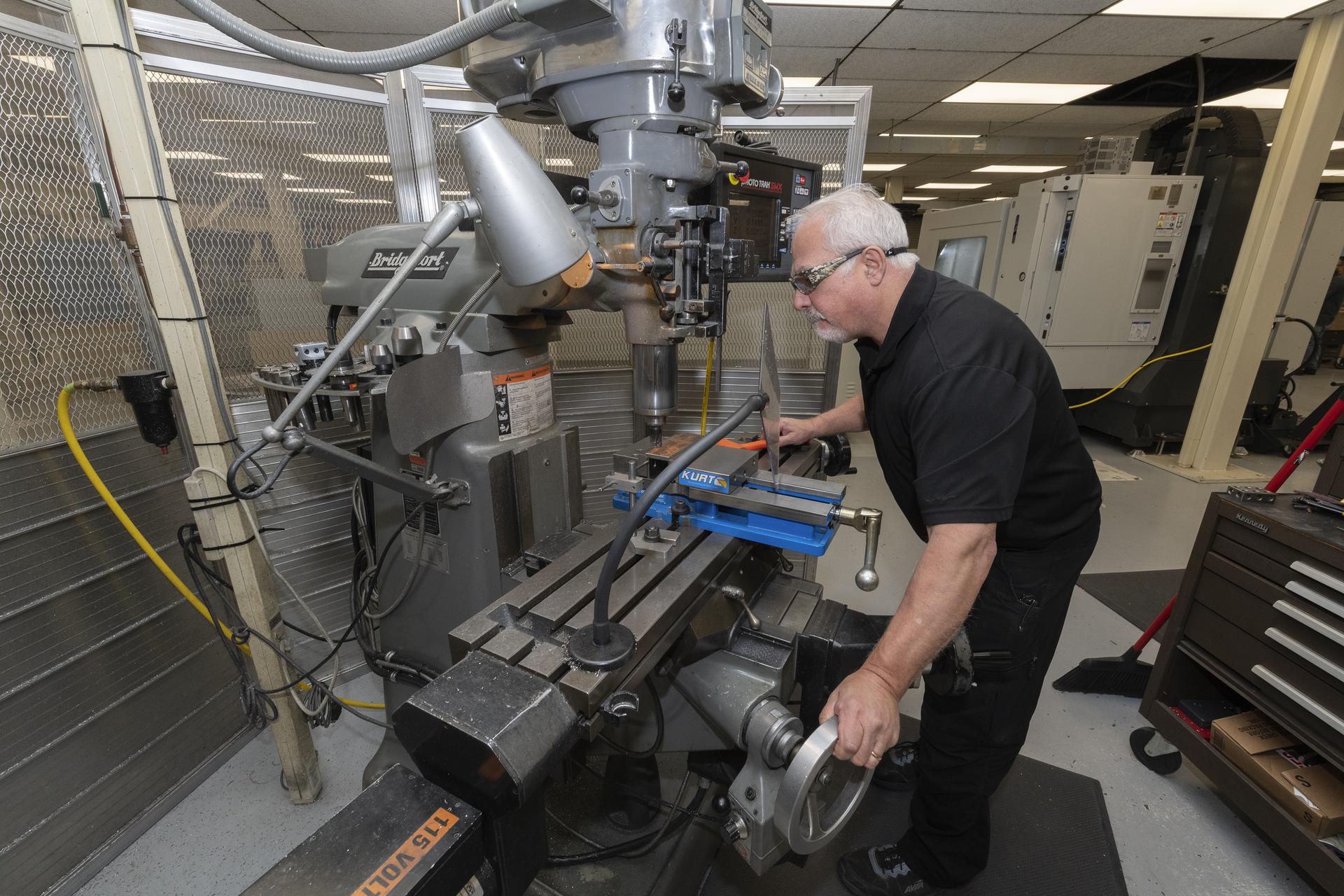 A man wearing all black and safety googles is working with a fabrication machine. He is holding a round wheel to operate a piece of the machine. In the background there are black tool benches and other tall machines.