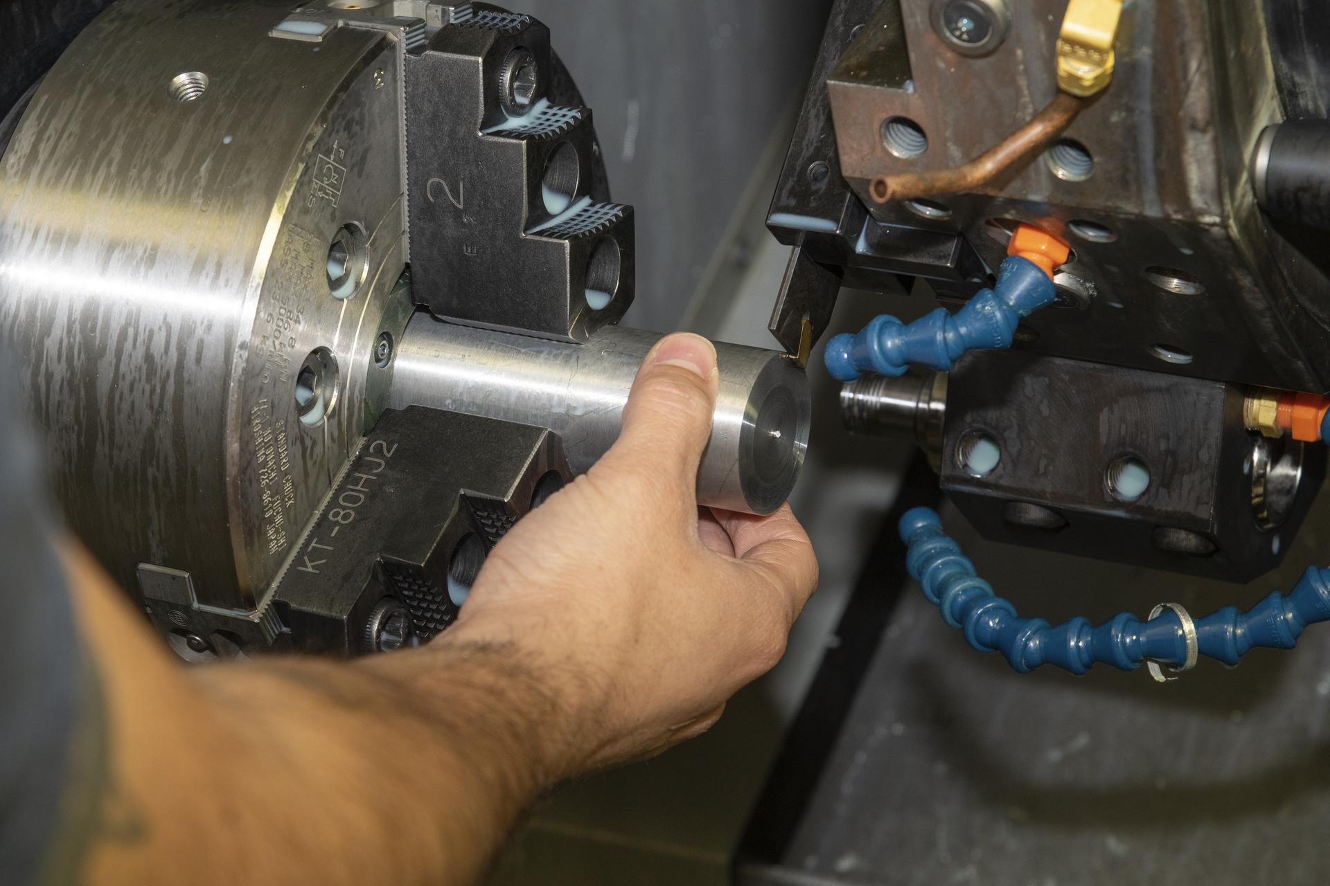 A person positions a cylindrical metal workpiece into the jaws of a lathe chuck. The shiny metal cylinder is partially inserted into the clamping mechanism with visible textured steel jaws and engraved markings. To the right, a cutting tool is mounted on a tooling block, surrounded by flexible blue coolant hoses with orange nozzles. The scene shows the close-up precision required in machining as the operator aligns the metal piece before cutting.