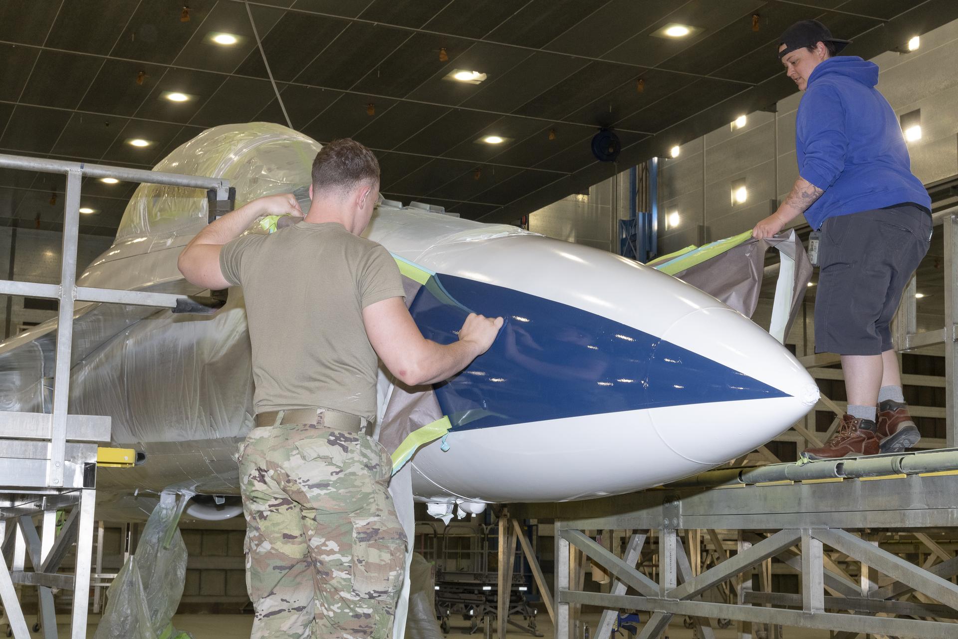 Two people remove masking off an aircraft. The white and blue painted nose is exposed.