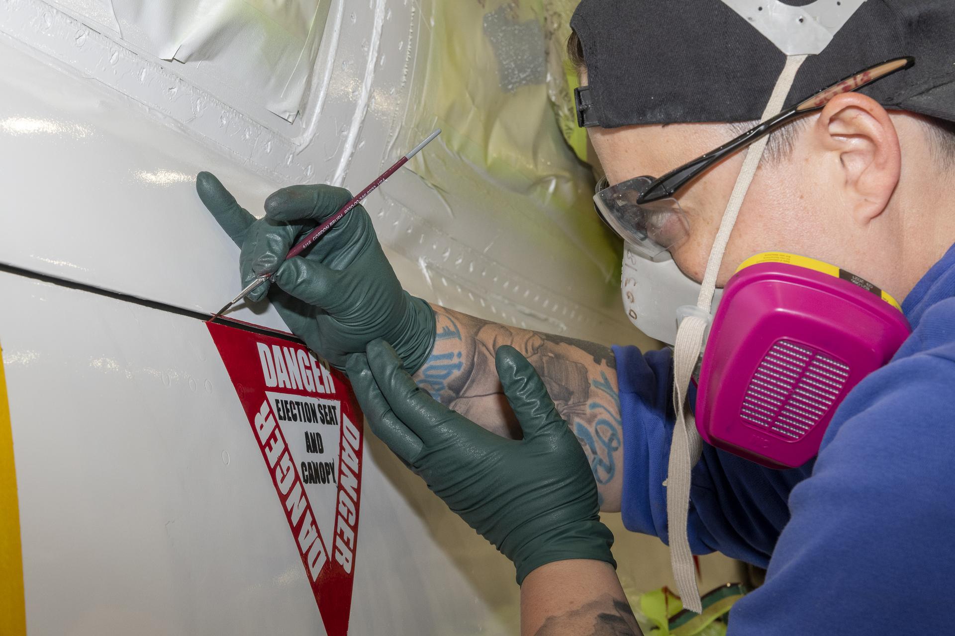 A person wearing pink, yellow, and gray protective head gear carefully paints a warning on the side of an aircraft using a thin brush.