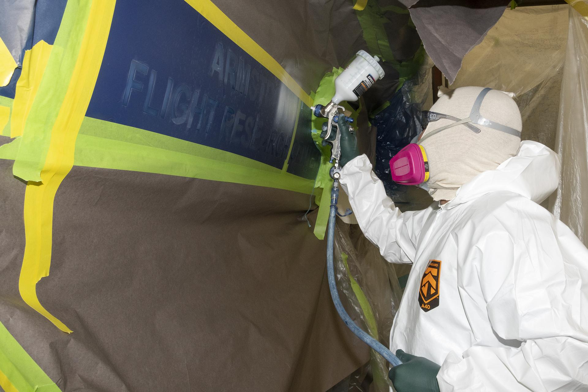 A person wearing a while protective suit and pink, yellow, and gray protective head gear paints over a stencil on the side of an aircraft.