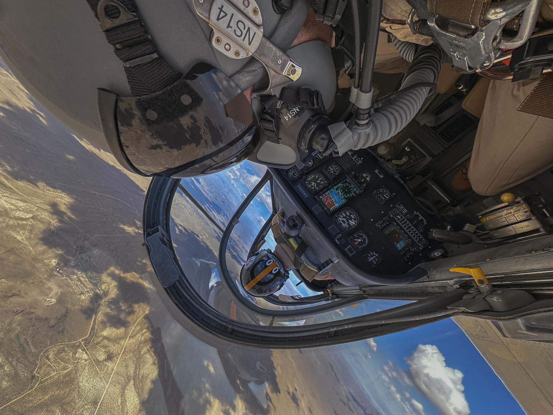 Inside of an aircraft cockpit is shown from the upside down perspective with two men in tan flight suits sitting inside. The side of one helmet, oxygen mask and visor is seen for one of the two men as well as controls inside the aircraft. The second helmet is seen from the back as the man sitting in the front is piloting the aircraft. You can see land below through the window of the aircraft.