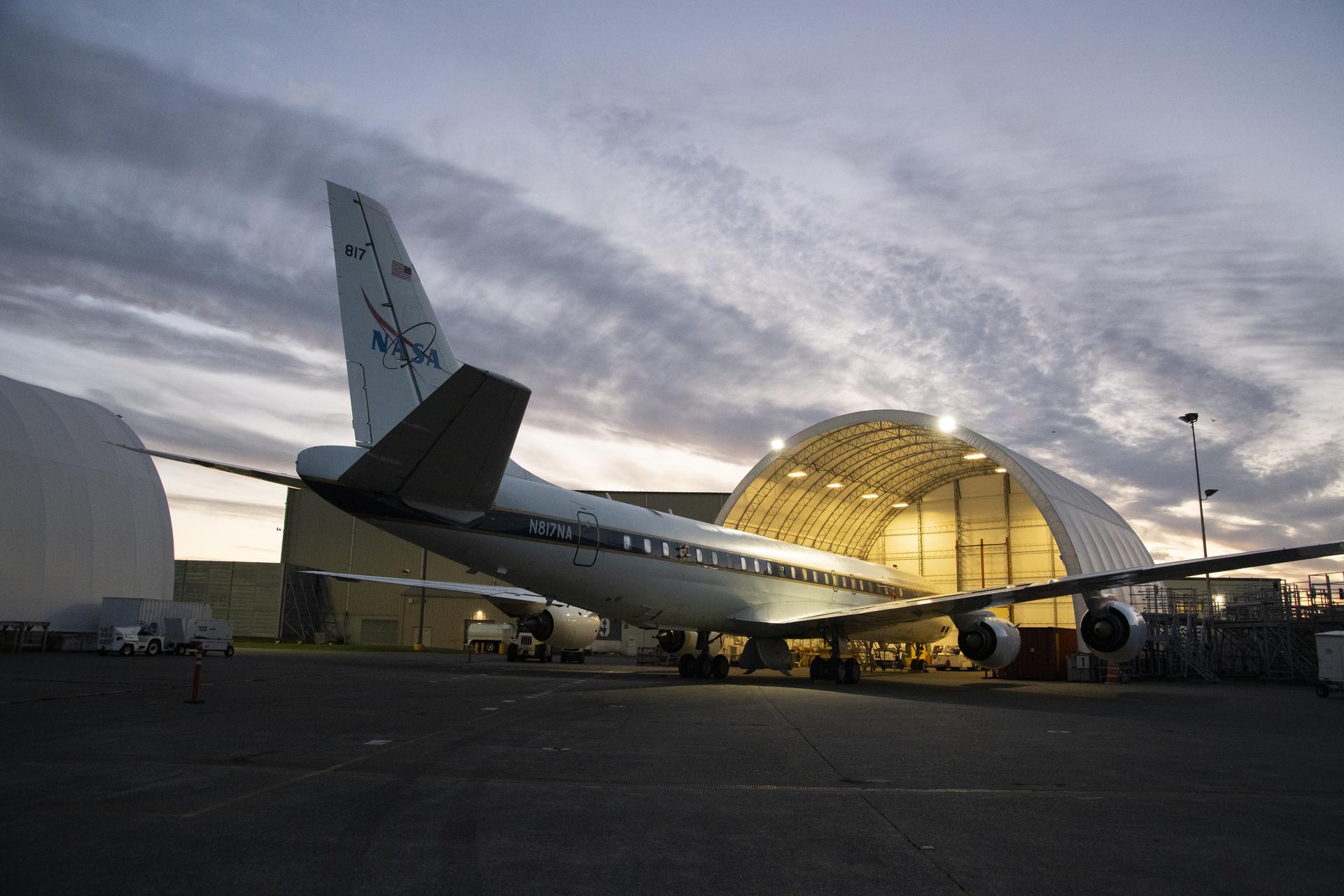 O avião DC-8 da NASA fica com o nariz dentro de um hangar à noite nas instalações de serviços de aviação onde foi alojado durante a campanha.