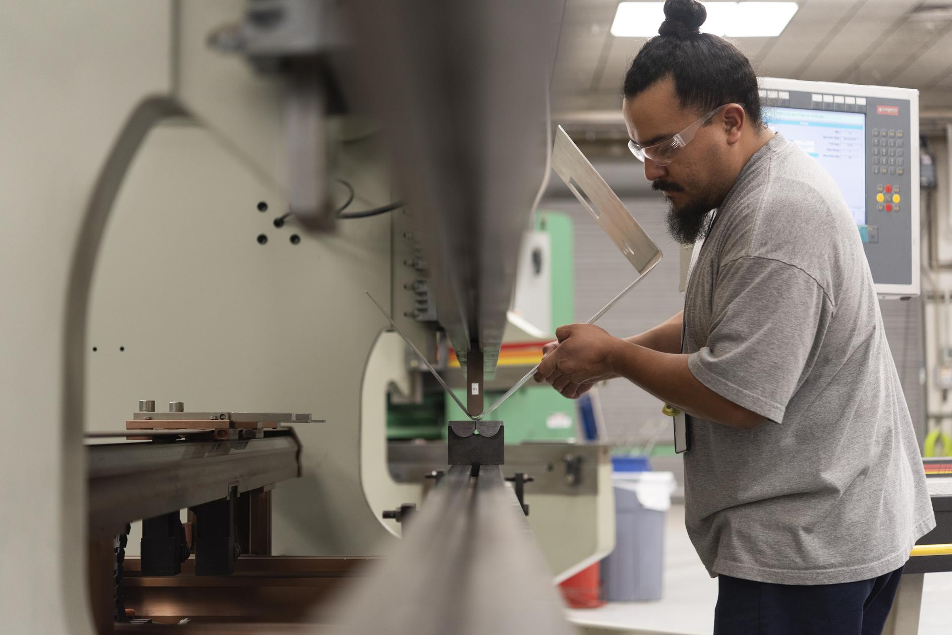 A technician stands at a large metal bending machine, carefully positioning a sheet of aluminum on the central die. Viewed from low angle along the machine’s long metal guide rail, the bending arm stretches into the foreground, emphasizing the scale of the equipment. The technician holds the sheet steady near the press brake while monitoring the alignment. Behind the workstation, a control panel with illuminated buttons and a digital display is visible, along with industrial shop equipment in the background.