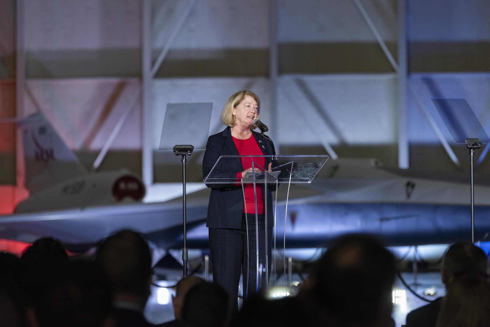 View of a lit stage inside a hangar. A woman speaks at a podium. In the immediate background, the X-59 sits, newly painted in its red, white, blue and gold livery.