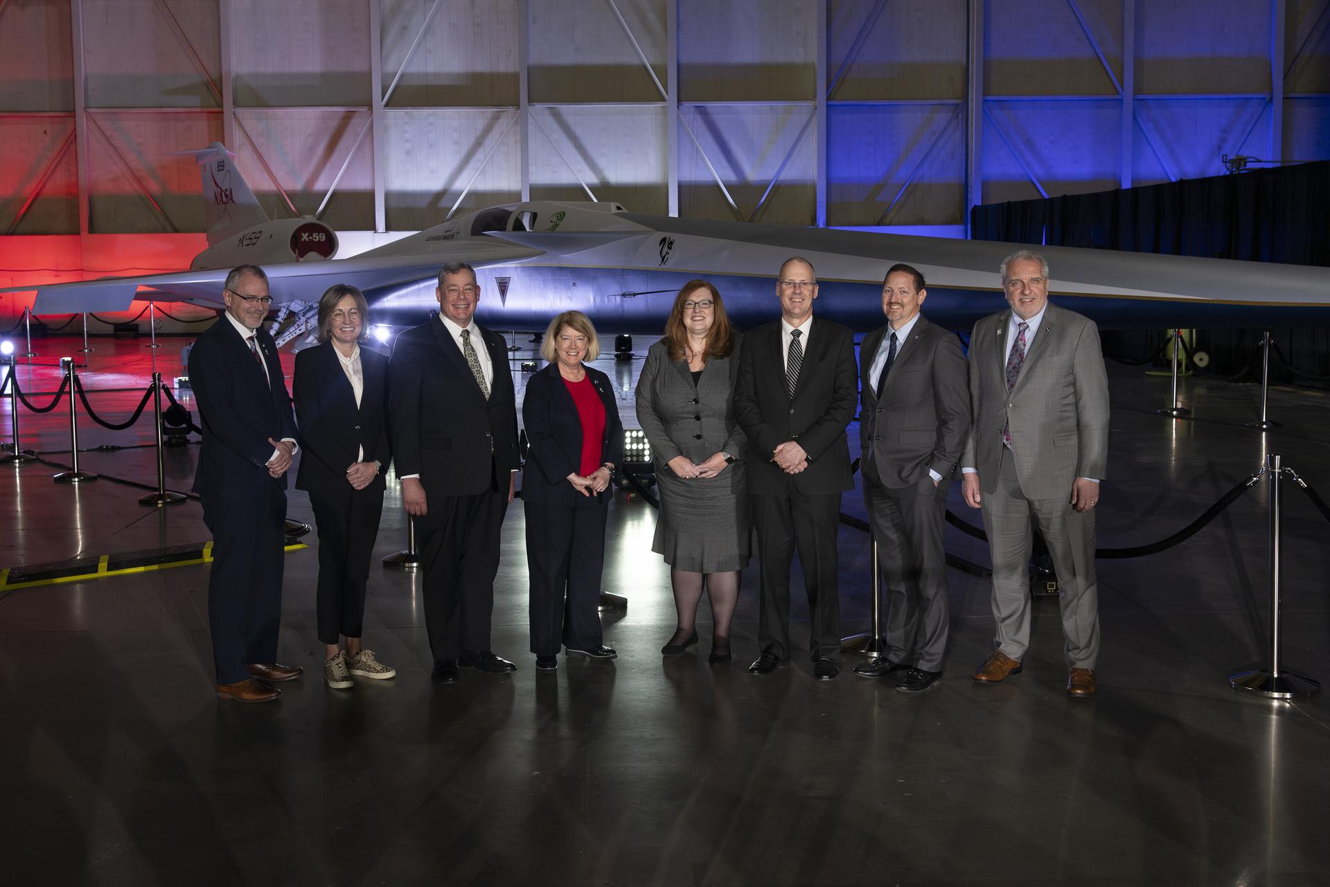 View of a lit stage inside a hangar. A group of men and women stand in front of the nose of the X-59, sitting immediately in the background, newly painted in its red, white, blue and gold livery.