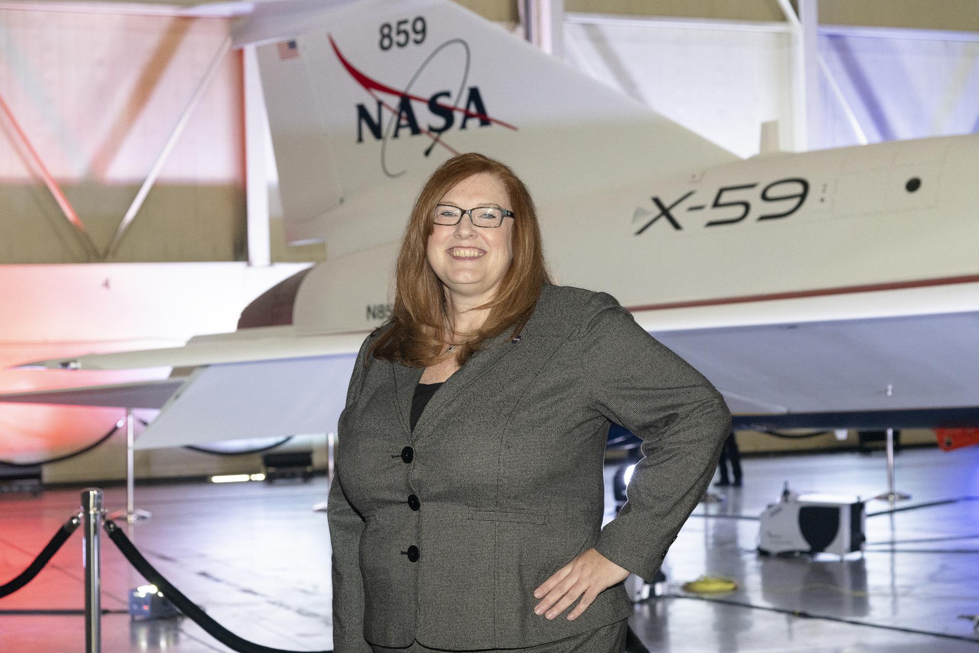 View inside a dimly lit hangar. A woman stands next to the tail of the X-59, facing the camera. In the immediate background, the X-59 sits, newly painted in its red, white, blue and gold livery.