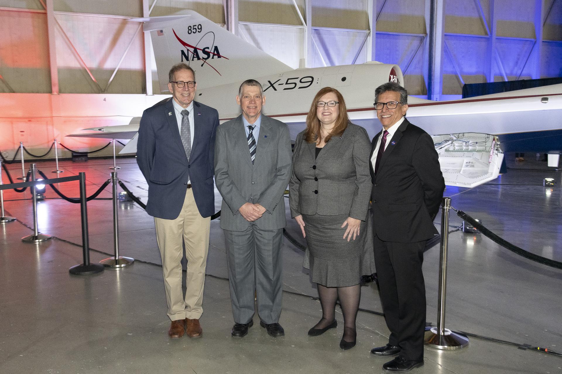 View inside a dimly lit hangar. A group consisting of men and women stand in front of the tail of the X-59, sitting immediately in the background, newly painted in its red, white, blue and gold livery.