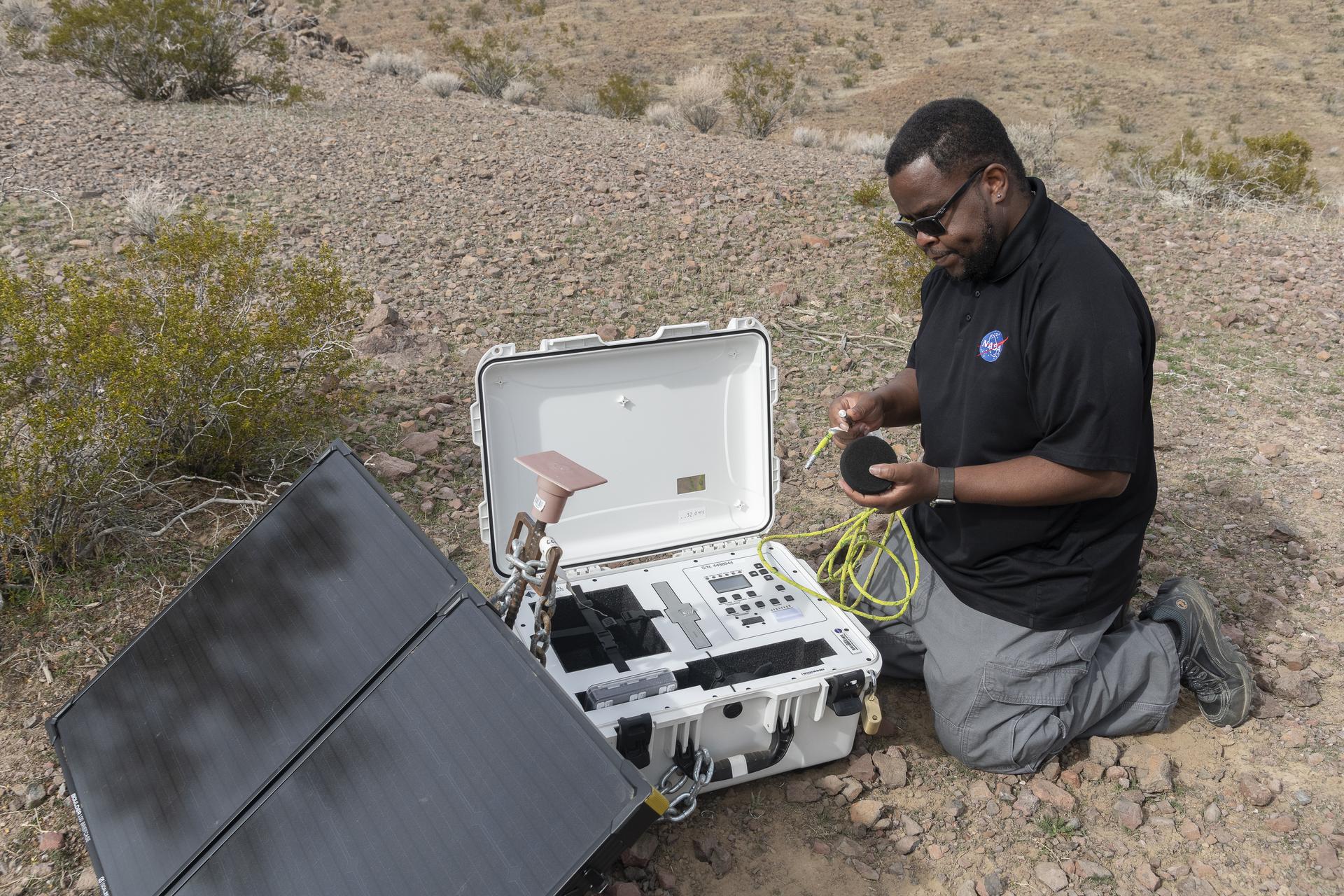 A man sets up recording equipment and a solar panel in the California desert.