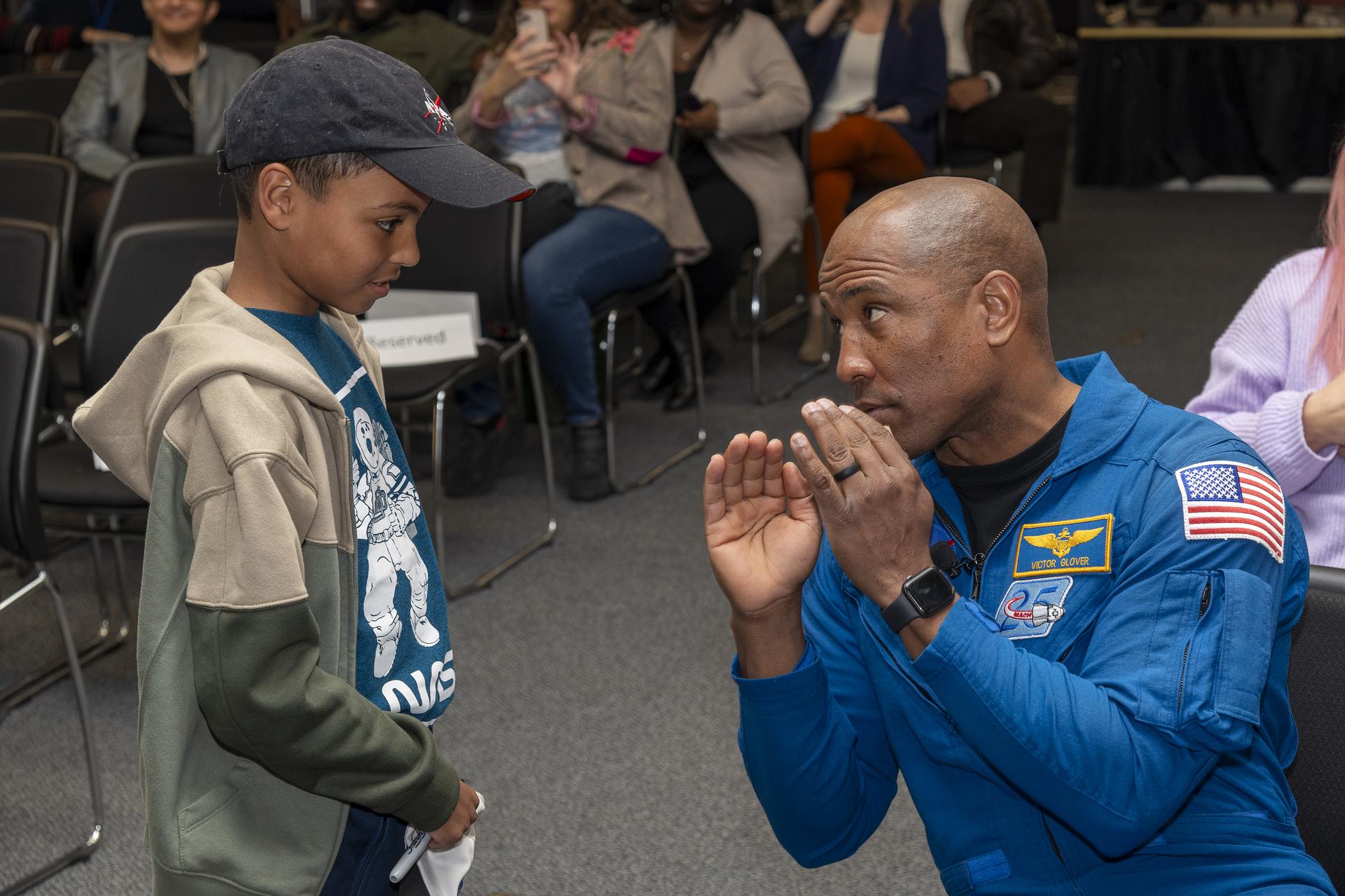 A black man and black child discuss, facing each other at ground level. The man gestures with his hands while wearing a bright blue flight suit with mission patches. The boy, in a green and tan jacket, teal shirt, and NASA baseball hat, listens intently. They are in an auditorium with chairs behind them.