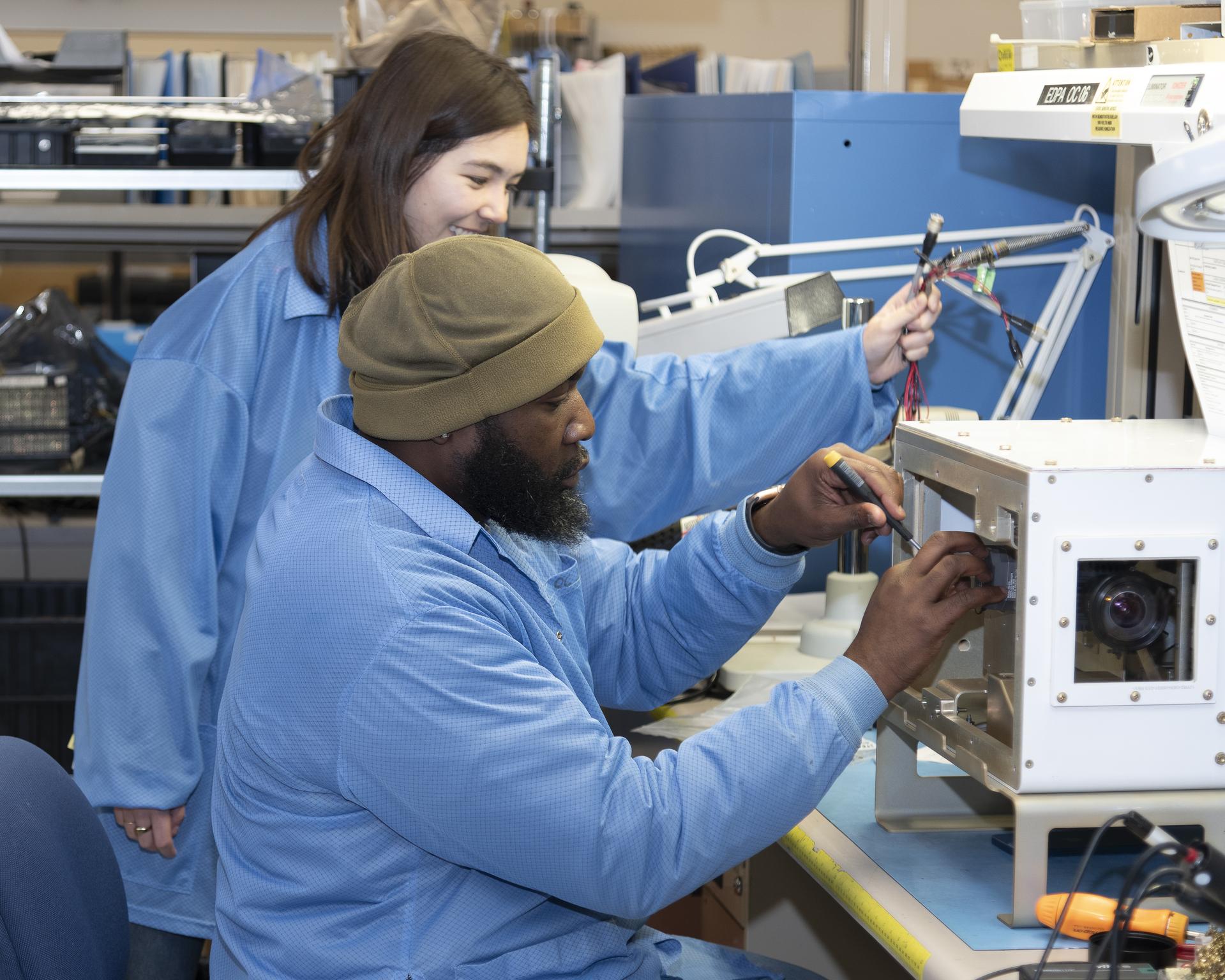 A man in the foreground and a woman in the background, both wearing light blue lab coats, install wiring, sensors, and cameras into a white cube that sits on a desk. The mean wears a tan winter hat. There is a metal shelf in the background full of hardware, wires, and baskets housing other lab equipment.