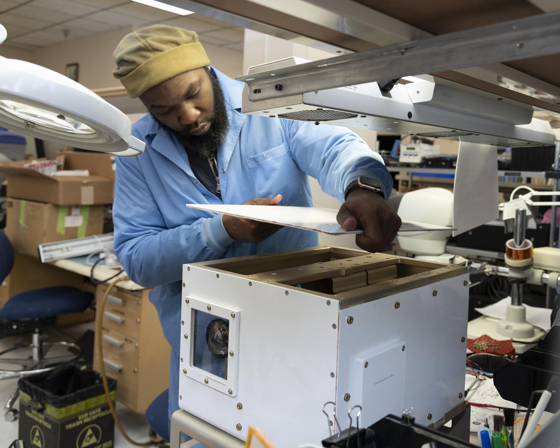 A man wearing a light blue lab coat inside of a lab installs the top back onto a white cube that sits on a desk. The man wears a tan winter hat. The background is filled with boxes, other lab equipment, a desk and rolling blue chair.