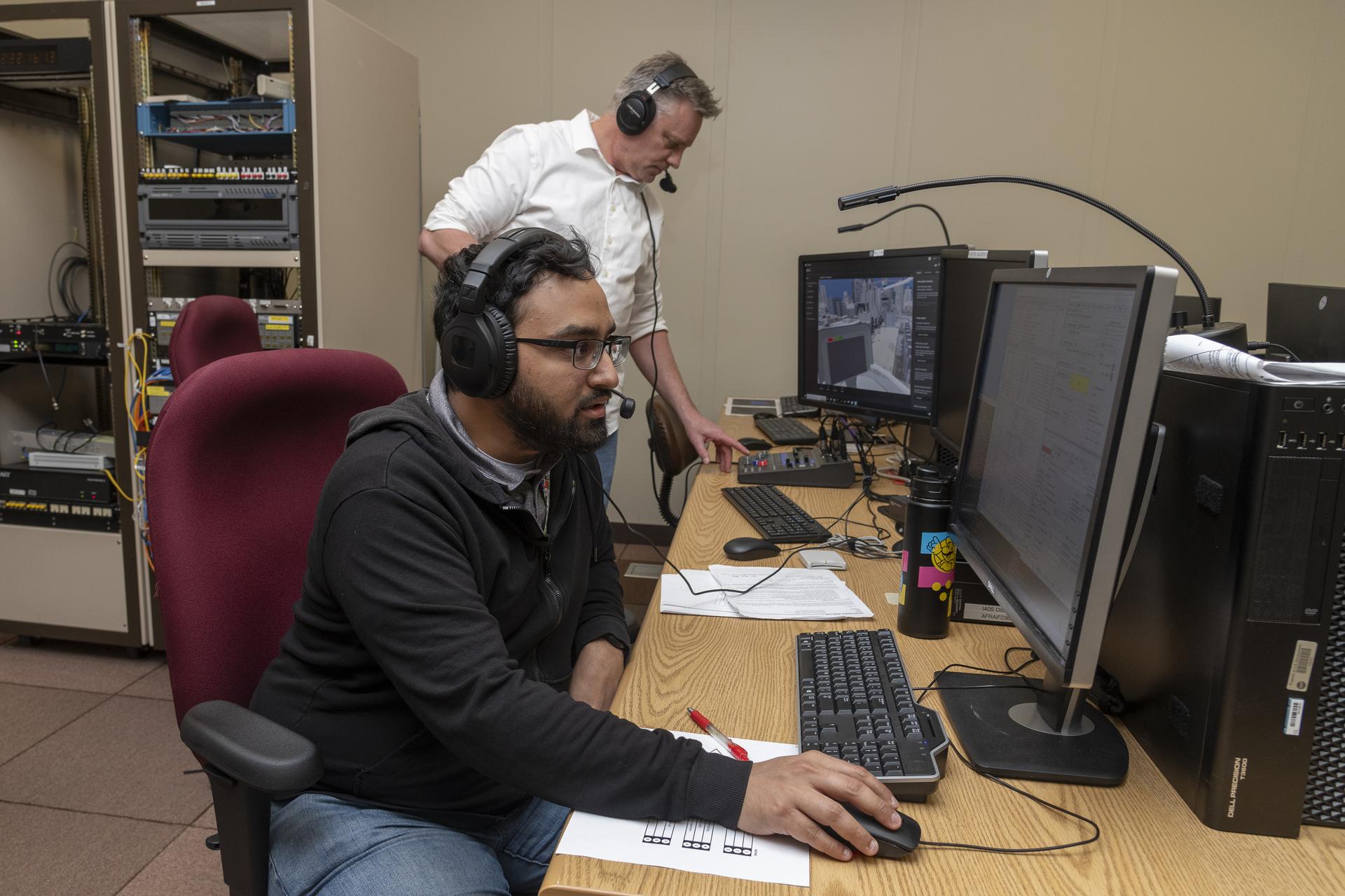 Two men sit in view in front of two computers with two microphones and with two keyboards. They both wear headsets with microphones. The man in the foreground is wearing glasses, a black hooded jacket and jeans. The man in the background is wearing a white button down shirt and jeans and pushes a button on a keyboard with one hand. On one screen, you can see a animated scene of the inside of an airplane cockpit and buildings below.