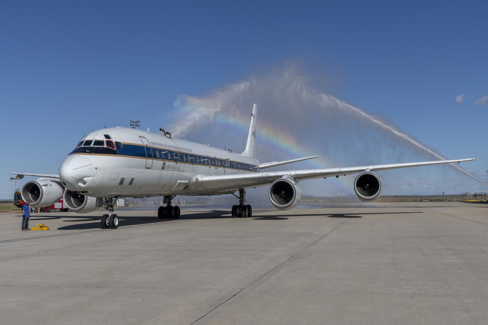 An aircraft taxis through a water salute streaming from water hoses from two firetrucks positioned on either side of the aircraft. The converging water streams create a rainbow above the aircraft as it passes under the arc of spraying water.