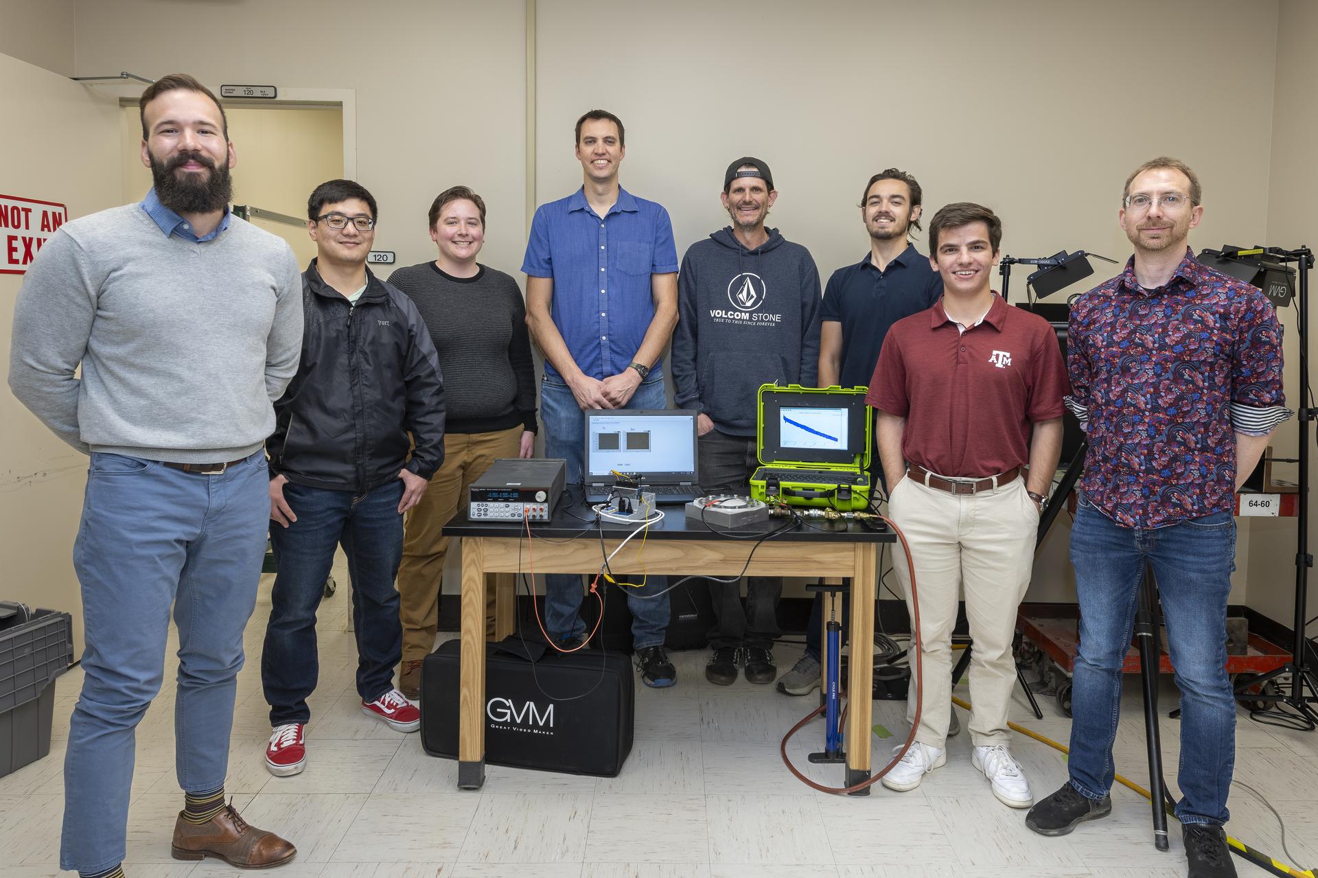 A group of people stand by a table that has a computer, instrumentation, and a round test fixture on it.