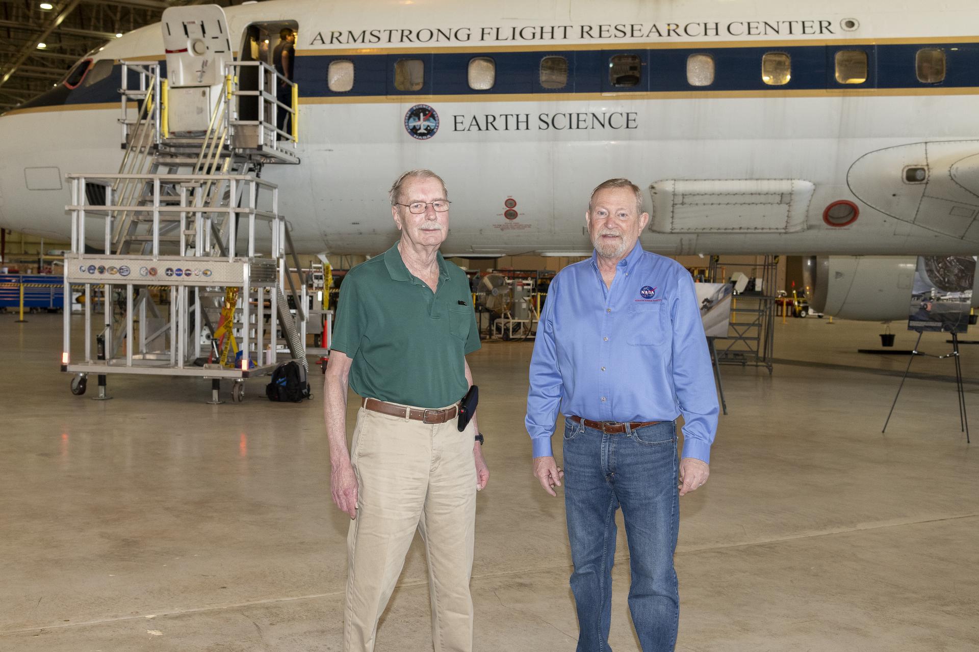 Two men stand in the foreground. A large aircraft is in the background.