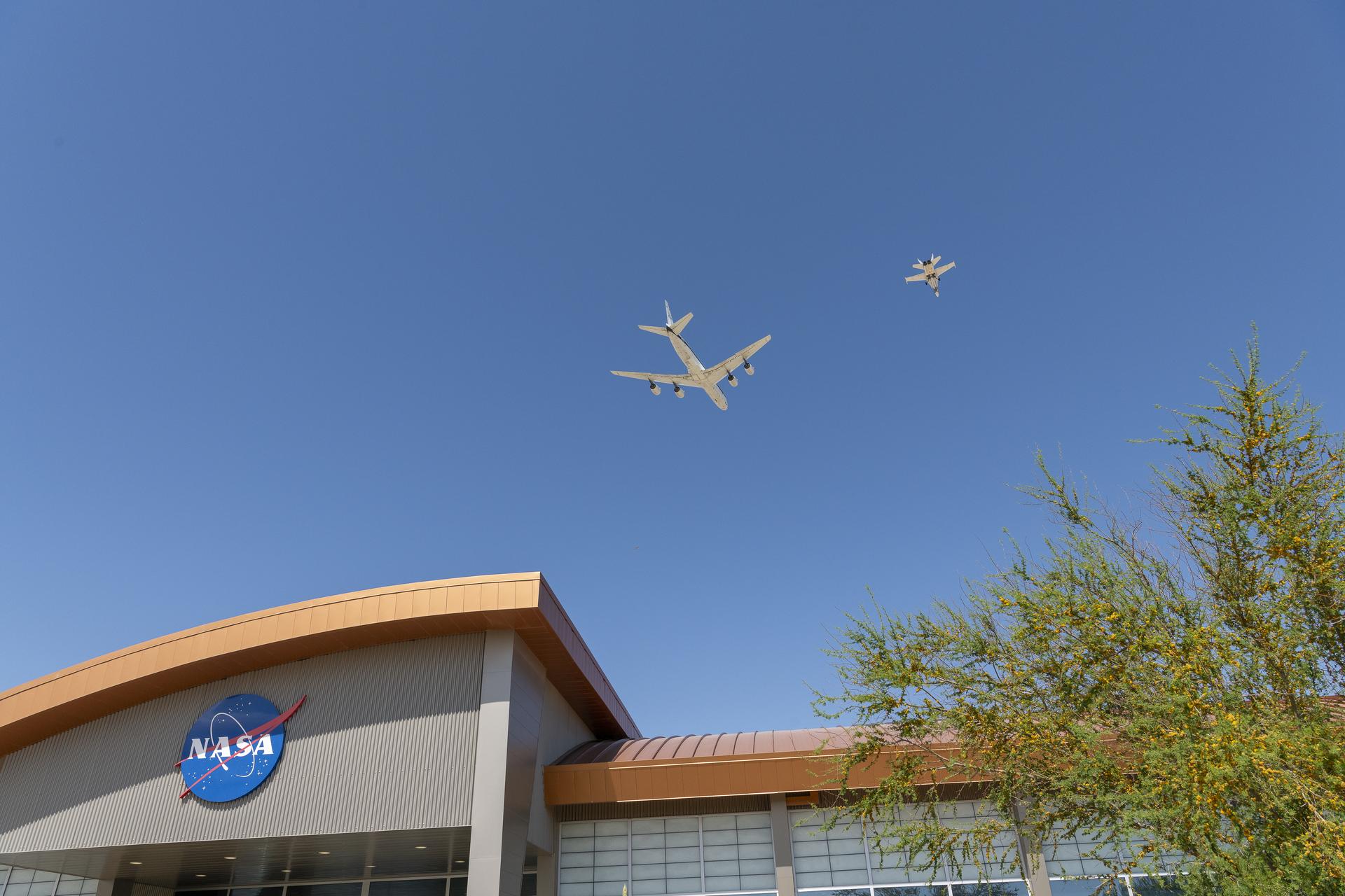 A large white plane and smaller chase plane fly over the top of a building with a NASA logo on the front. A sunny blue sky is behind the building and planes.