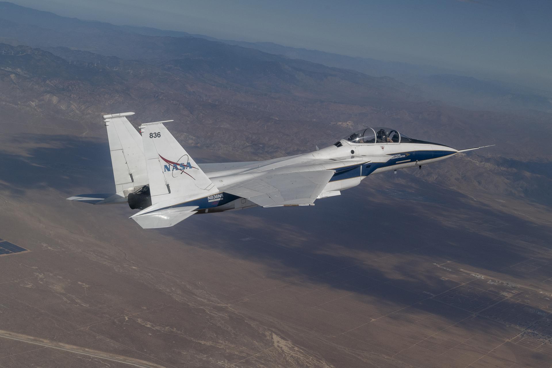 An F-15B aircraft, painted white and blue, is shown in flight with clouds above. The Shock Sensing Probe mounted to its nose during an Aug. 6, 2024, calibration flight over Edwards, California.