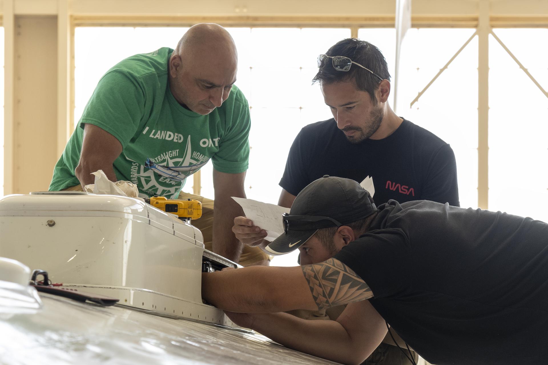 Three men examine an instrument: the man on the left has a green shirt and looks over the shoulder of the center man, who wears a black shirt and reads a piece of paper he holds in his hands. The man on the right is wearing a black shirt and cap, and uses his hands to engage with the inside of the white instrument body.