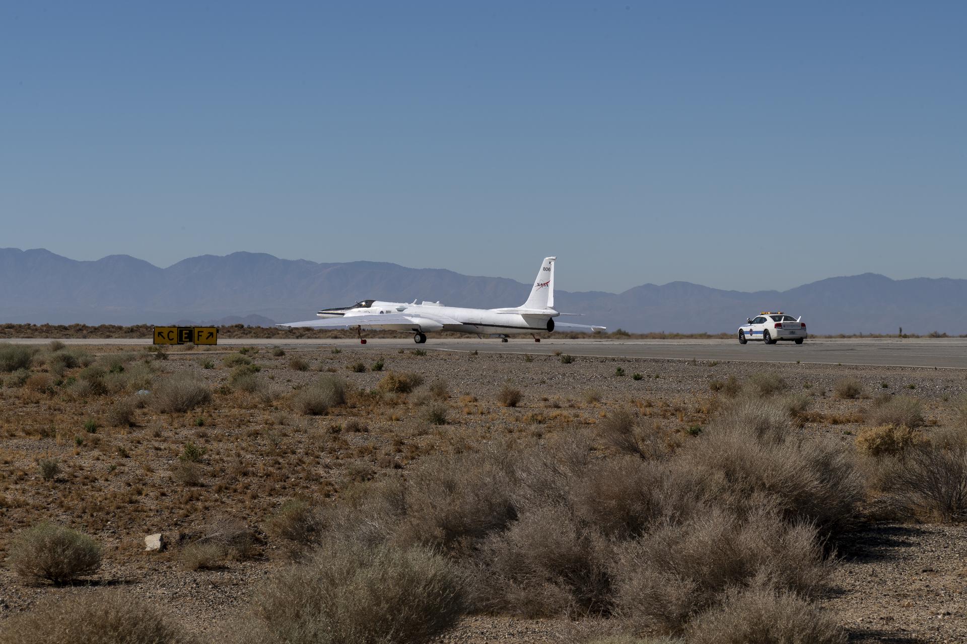 A white plane heads down a runway preparing for takeoff with a white chase vehicle following behind. The background is a clear, sunny day and the foreground is desert foliage and landscape.