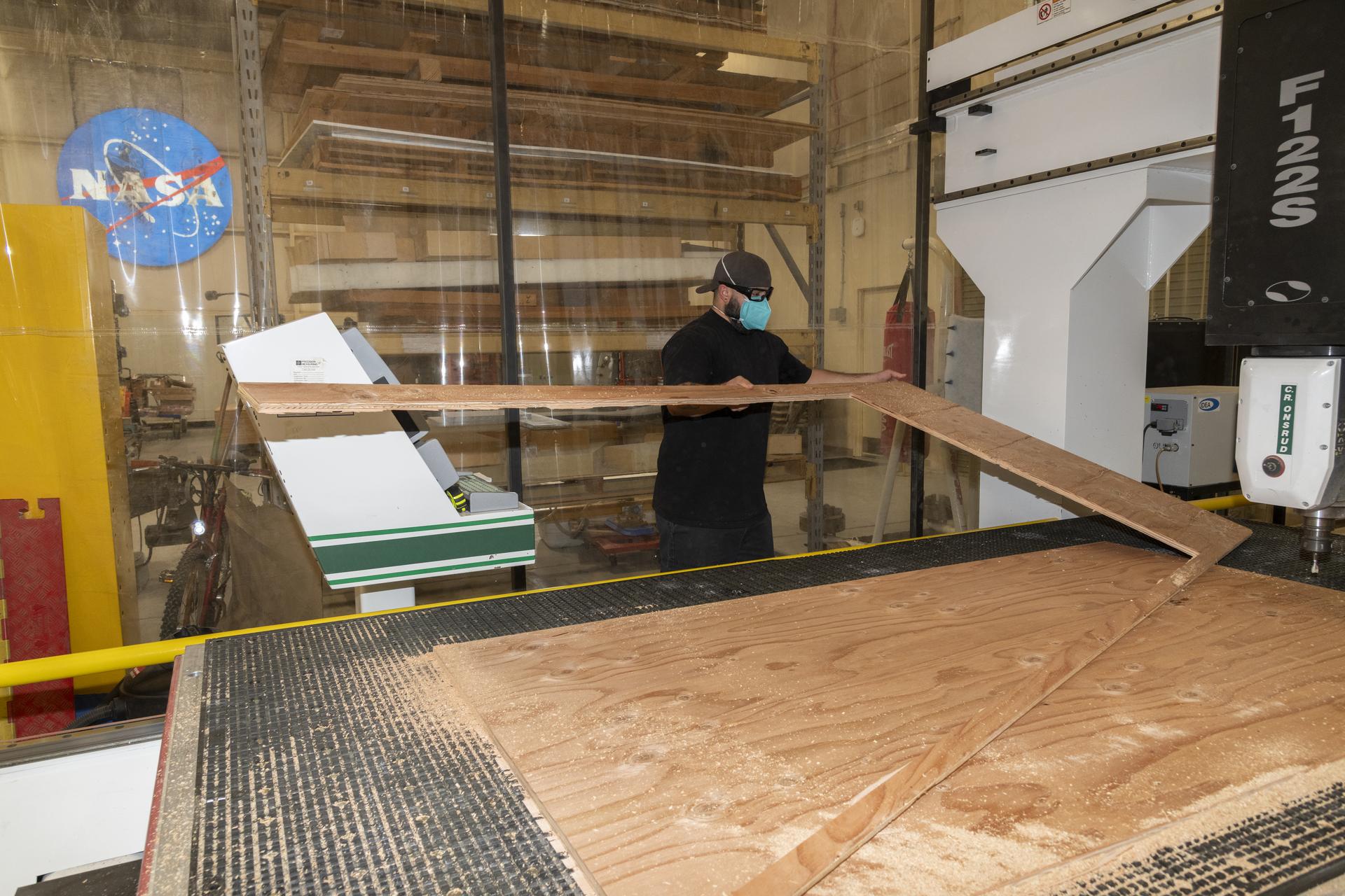 A man wearing a black shirt, a mask, glasses, and a baseball cap backwards handles part of a piece of light brown plywood as he is pulling it off a machine. There is clear plastic hanging from the wall behind him. The NASA logo is also hanging on the wall behind the plastic. There is wood dust around the area of the machine.