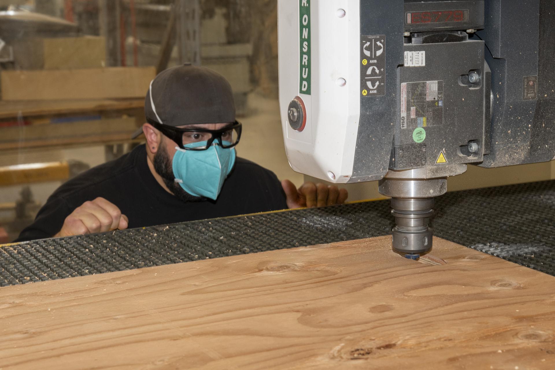 A man wearing a mask, glasses, and a baseball cap backwards watches as a silver drill-like machine cuts holes into plywood.