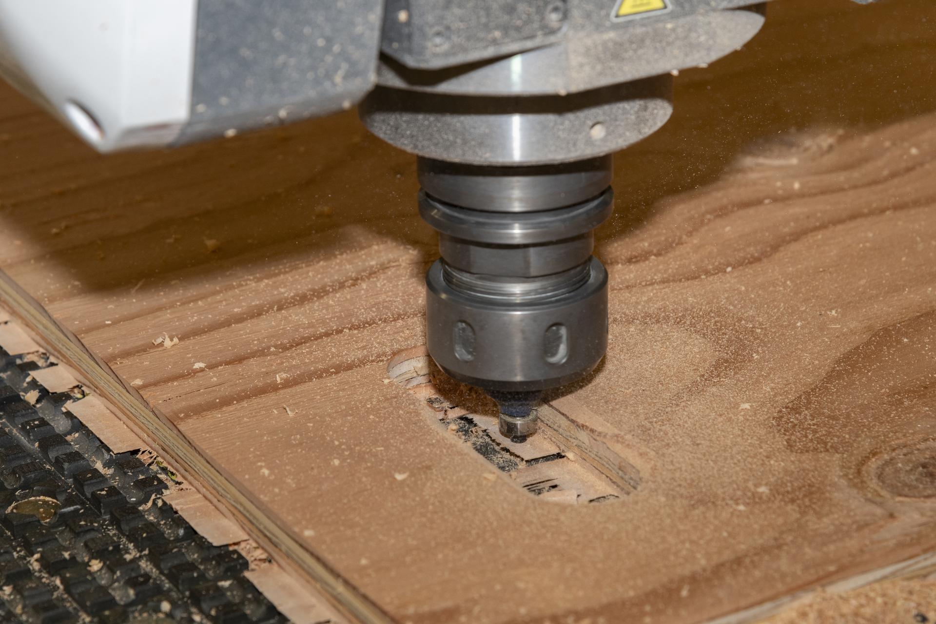 A silver drill-like machine cuts holes in light brown plywood. There are specks of wood dust around the cut.