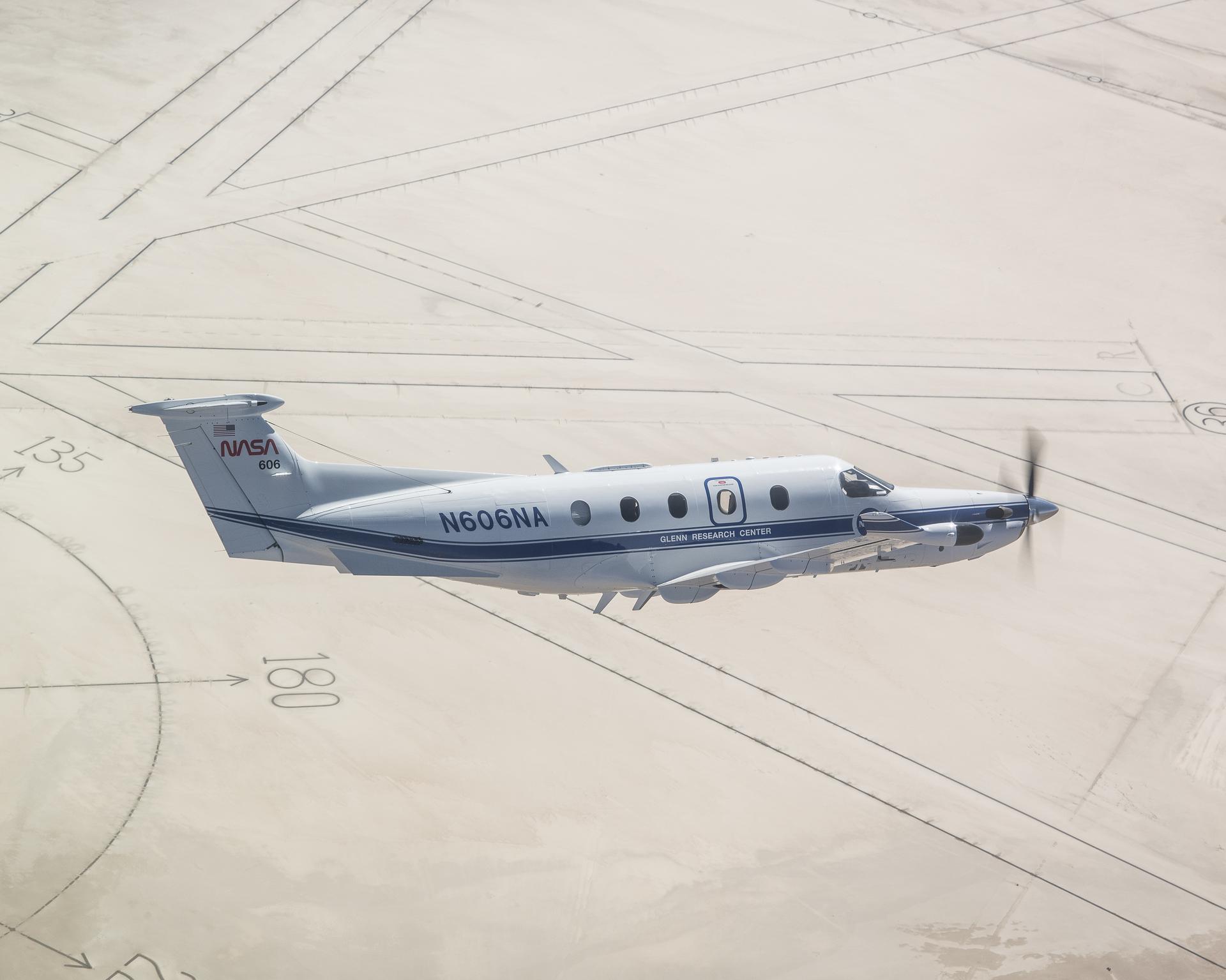 The tan sands of Rogers Dry Lakebed and the darkened directional lines of the compass rose at NASA’s Armstrong Flight Research Center in Edwards, California frame NASA’s newly painted Pilatus PC-12 airplane. The gleaming white plane with a blue stripe and blue N606NA number across the fuselage and NASA red worm logo on the tail, cuts across the clear, bright desert skies.