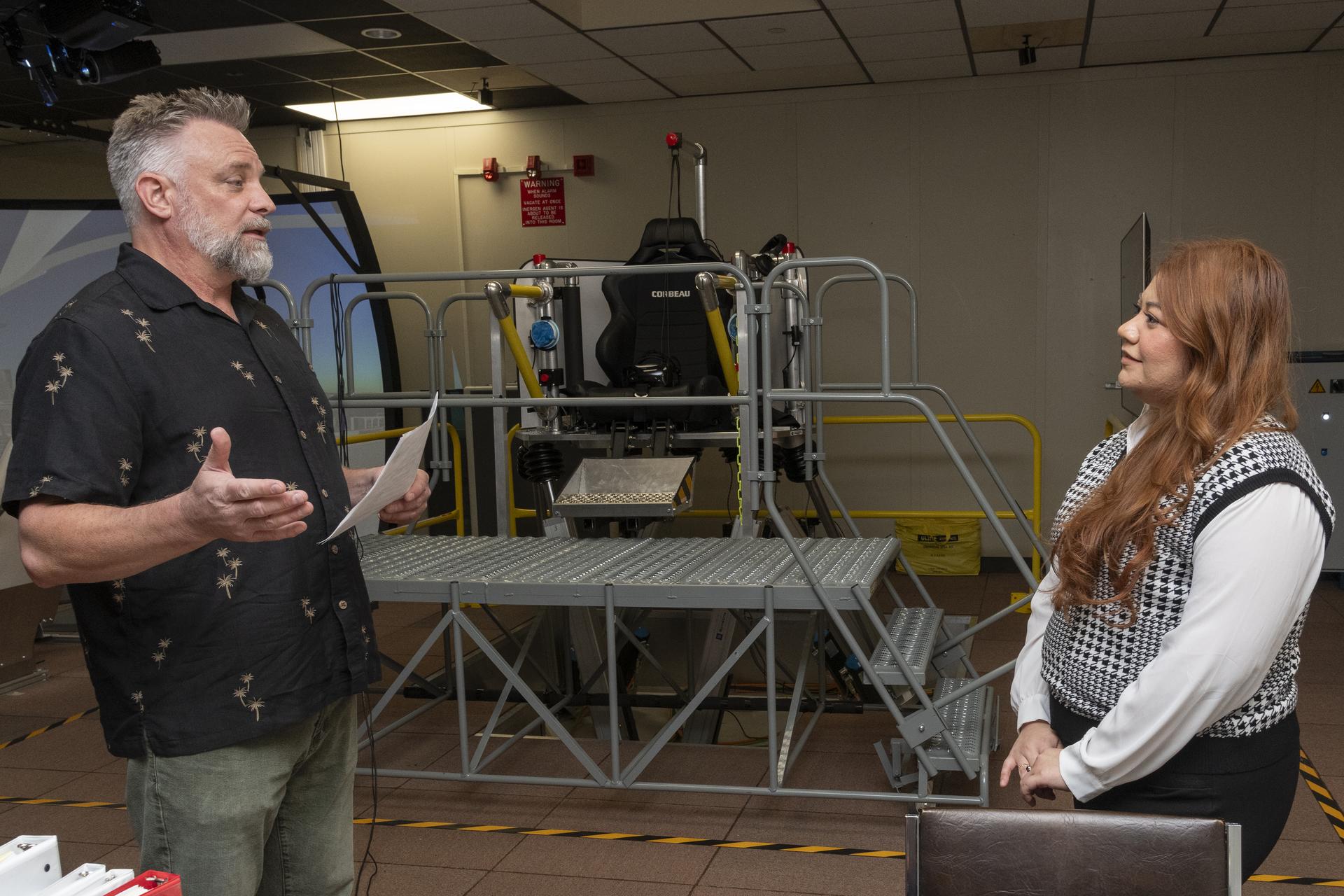 A man wearing a black shirt, standing on the left side of the image, faces a woman wearing a black and white vest on the right side of the image. The man holds a piece of paper in one hand and his other hand is stretched out to the side. Behind them sits a metal platform with steps, which houses a black seat.