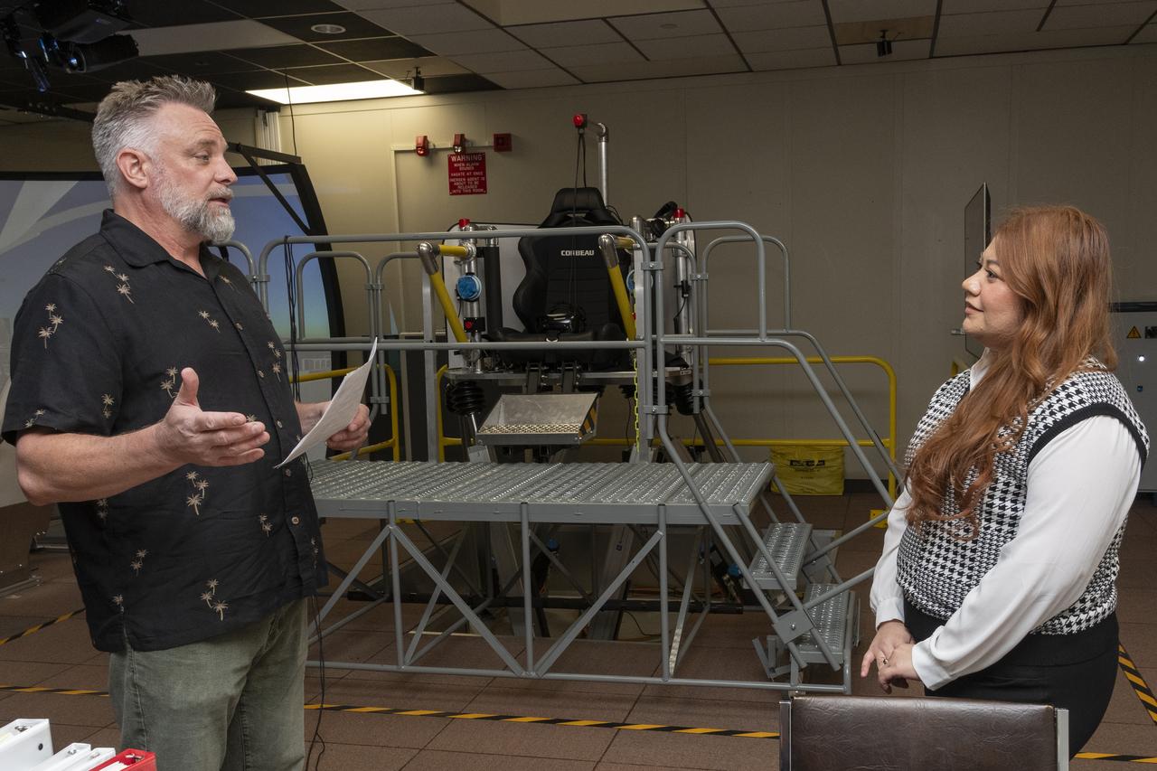 Curt Hanson, senior flight controls researcher for the Revolutionary Vertical Lift Technology project based at NASA’s Armstrong Flight Research Center in Edwards, California, explains the study about to begin to NASA employee and test subject Naomi Torres on Oct. 23, 2024. Behind them is the air taxi passenger ride quality simulator in NASA Armstrong’s Ride Quality Laboratory. Studies continue to better understand passenger comfort for future air taxi rides.