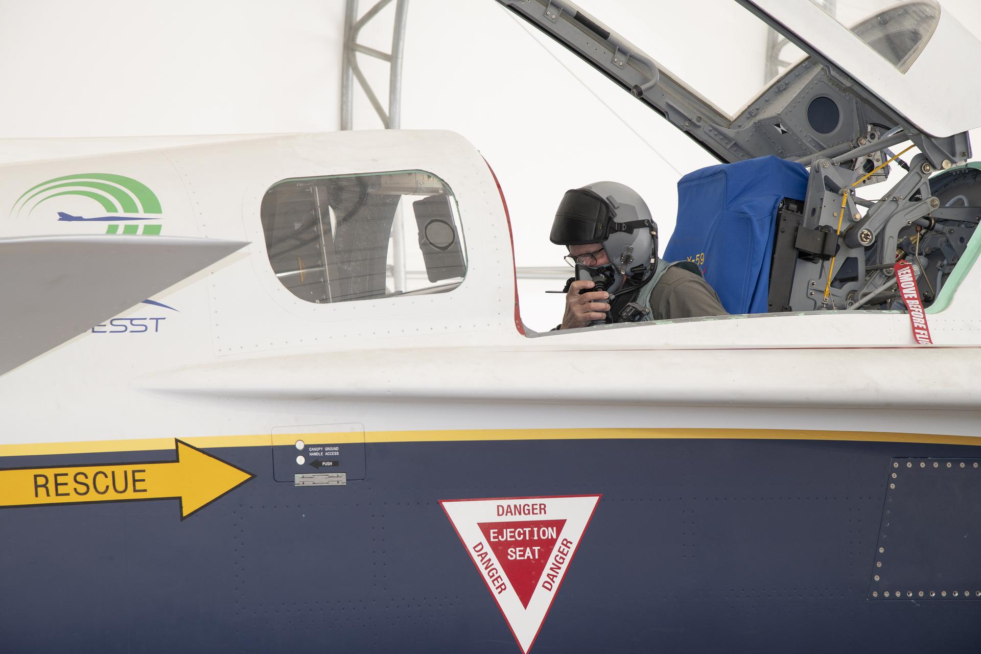 View of an aircraft cockpit inside a run stall. The canopy atop the cockpit is open. Inside, a pilot looks at his instruments as he wears a helmet and oxygen mask. The white, gold and blue paint scheme of the NASA aircraft is prominent.