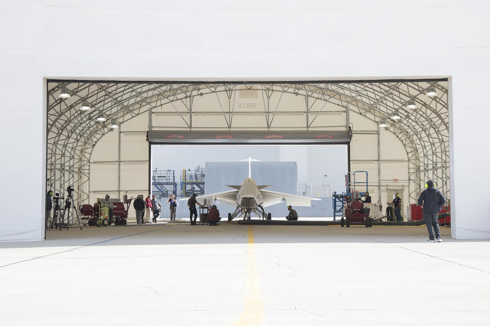 Direct view of the front of an aircraft, mostly in the shade of a run stall. Flaps on both wings are activated, moving downward. Team members wear protective ear guards and look at the aircraft from both sides.