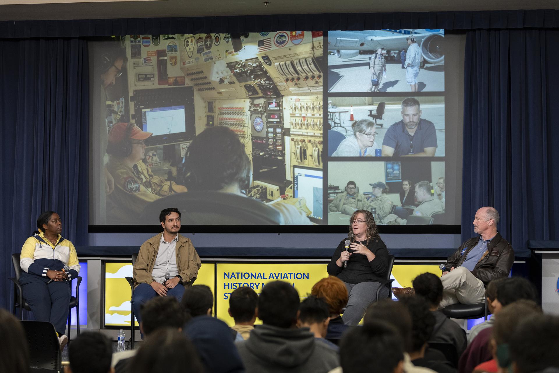 A group of four people are on a stage looking out and talking with students. Behind them is a presentation display of various images.
