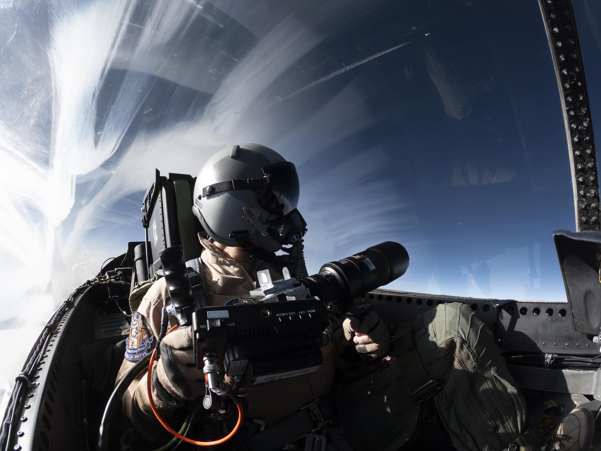 A NASA photographer sits in the rear seat of a research aircraft in flight, wearing a gray helmet with visor down and an oxygen mask. They hold a high-speed camera with a long lens on their lap, aimed out the left side of the canopy to capture schlieren images through the left side window of the canopy. The sky behind the photographer is deep blue on the right side, with flashes of white on the left side.