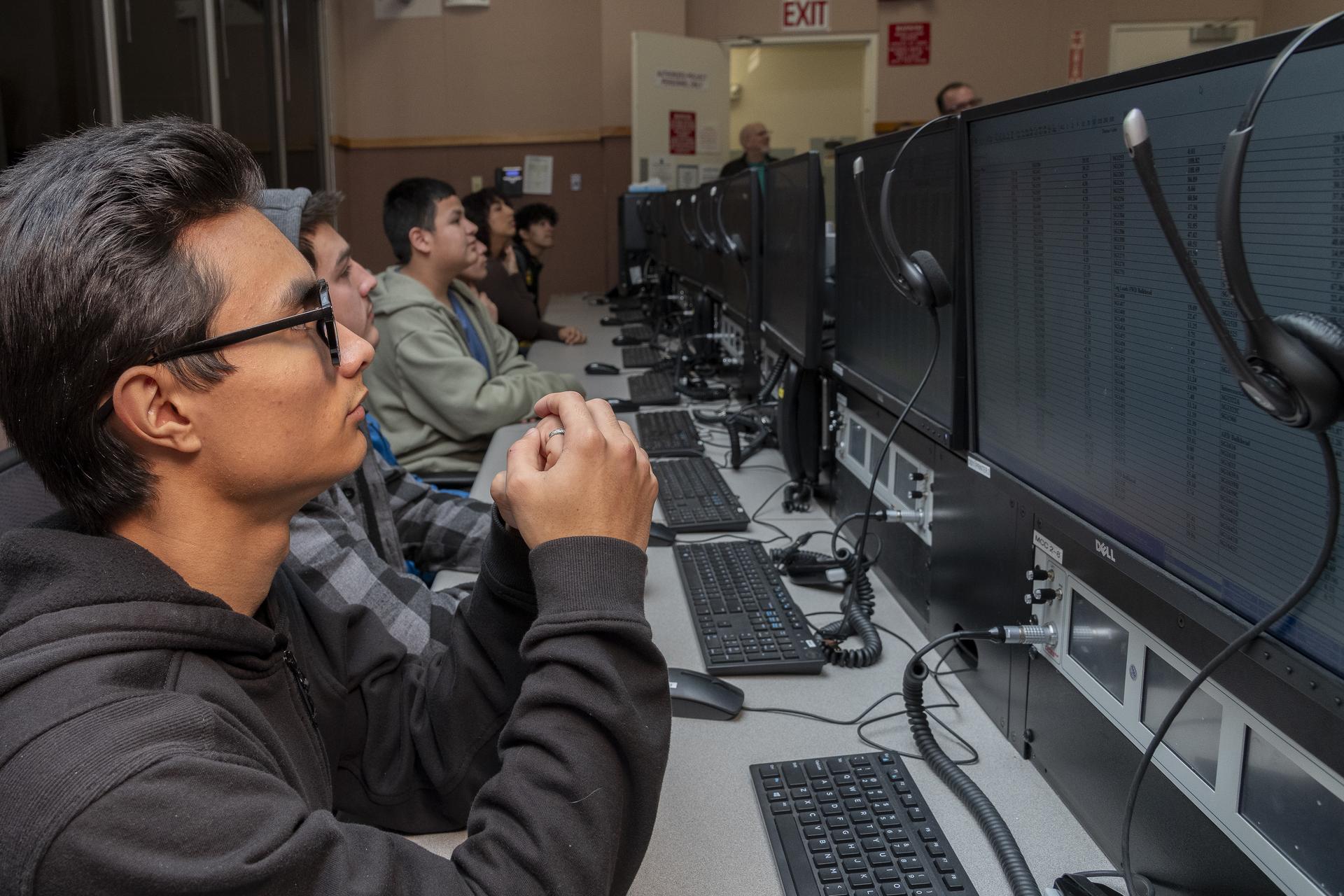 Students are sitting in a mission control room watching screens in front of them.