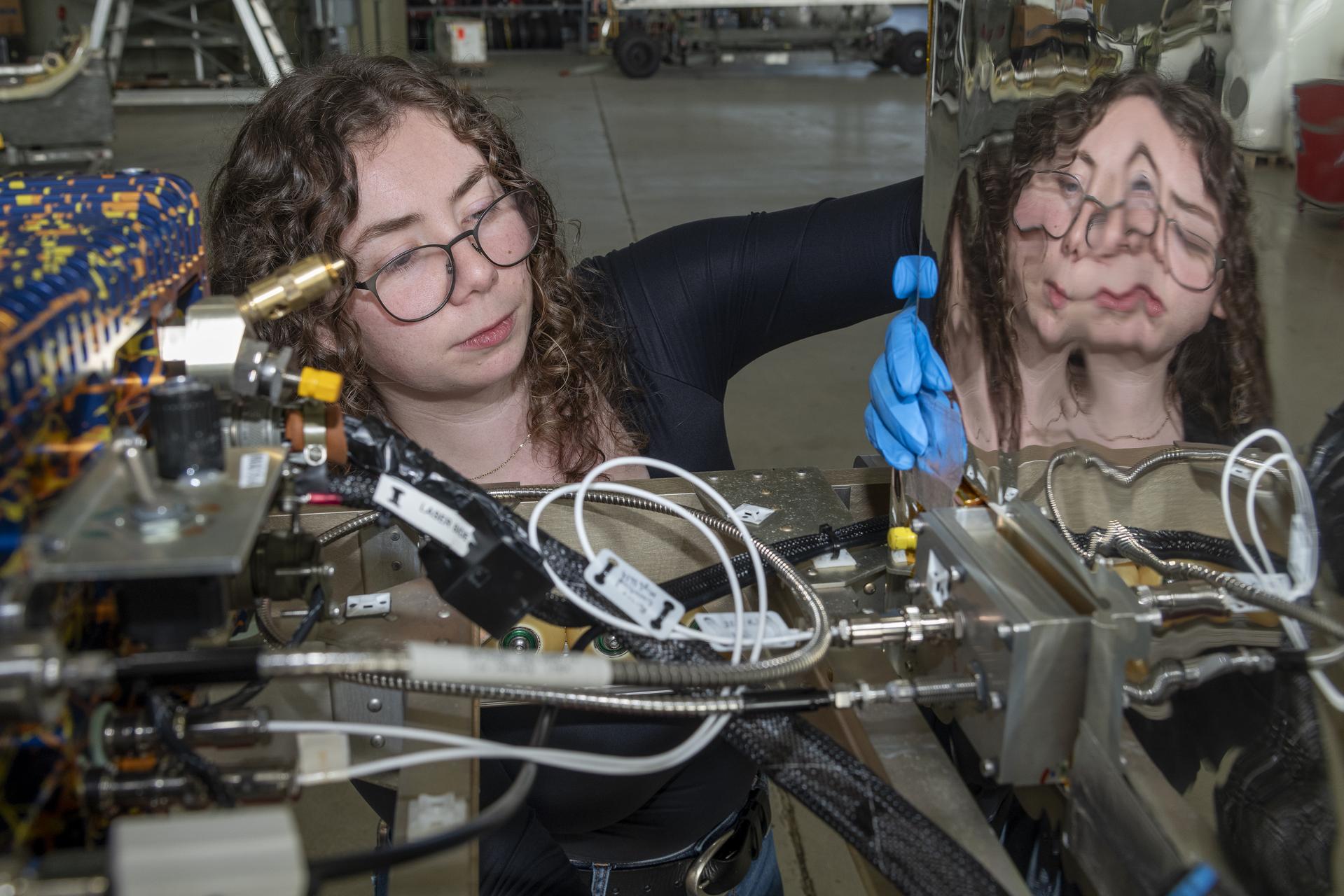 A woman face peers between two frames of a scientific instrument. The instrument is riddled with cables and cords and she manipulates some unseen component with blue rubber gloves. The right part of the instrument has a beveled reflective surface parallel to her face, and her reflection is distorted on the exposed metallic surface.