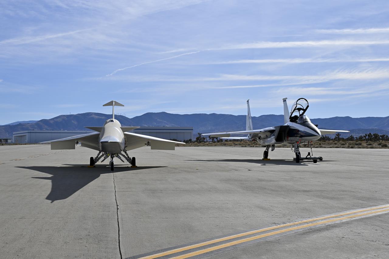 A NASA F-15 aircraft sits 20 feet off the left side of the X-59 aircraft, with a white hangar and hills in the background, during electromagnetic interference testing.