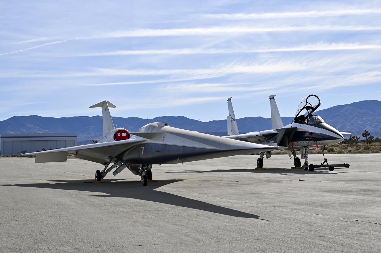 A NASA F-15 aircraft sits 20 feet off the left side of the X-59 aircraft, with a white hangar and hills in the background, during electromagnetic interference testing.