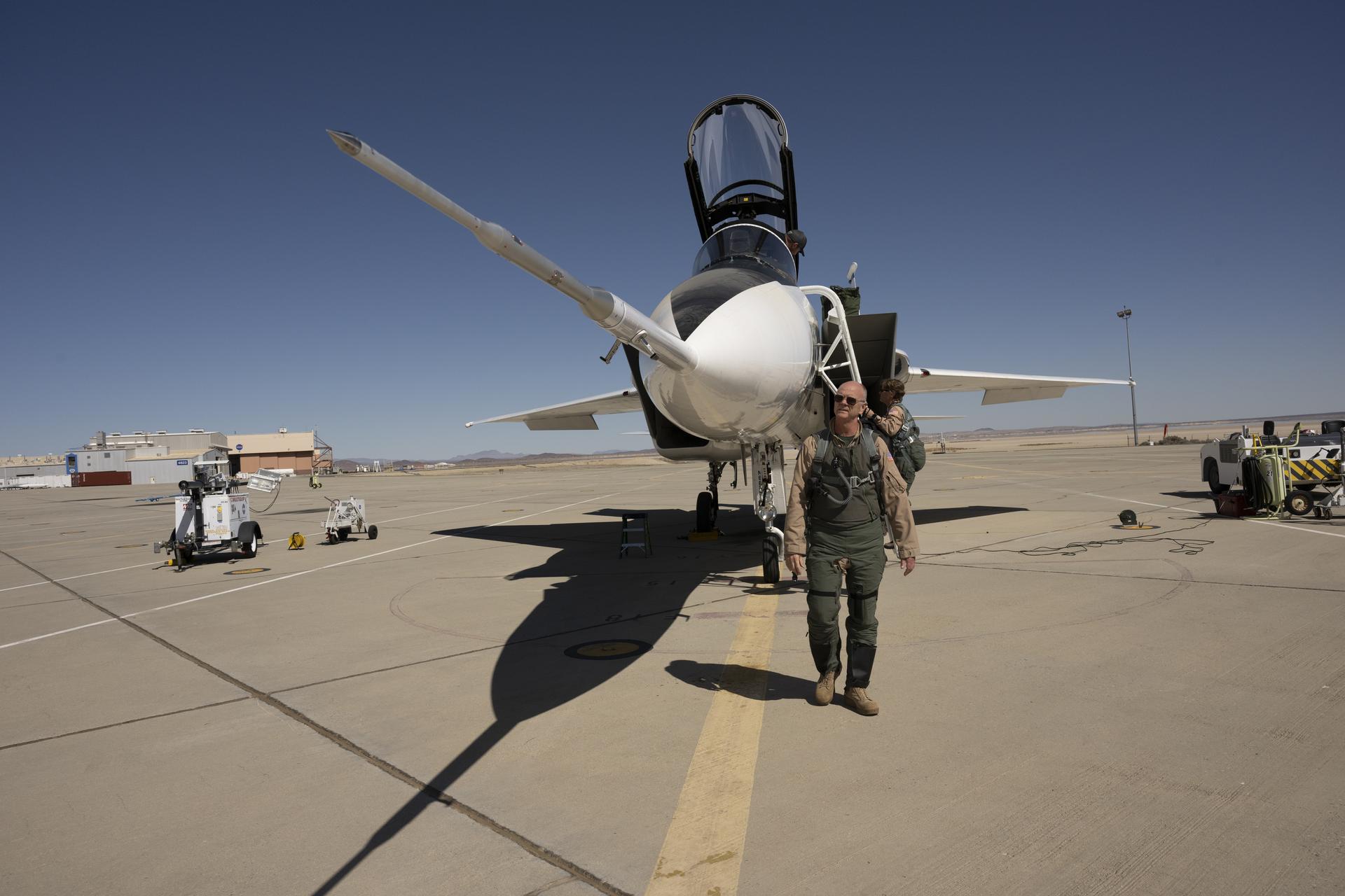 NASA test pilot Nils Larson, wearing a green flight suit and sunglasses, stands at the front of the agency’s F-15D research aircraft, looking up at a long, newly installed shock-sensing probe mounted on the nose. The aircraft is parked on the flightline at NASA’s Armstrong Flight Research Center in Edwards, California, with its canopy open and ladder down.